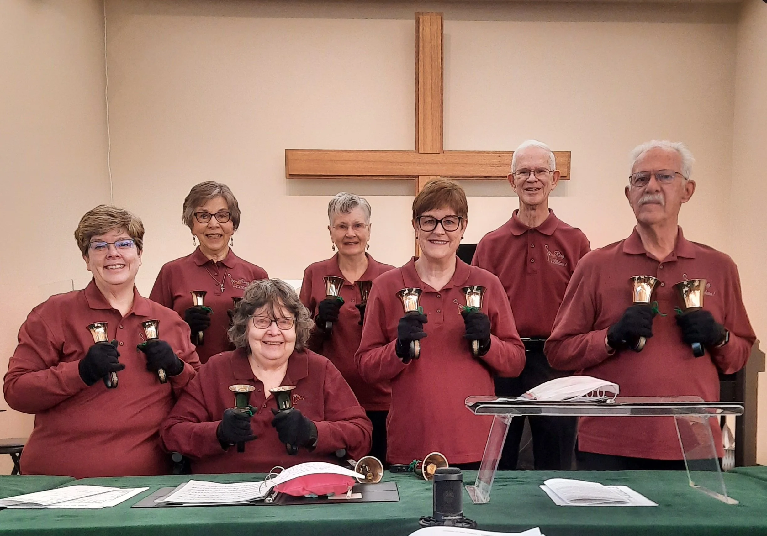 Group of seven older adults standing inside church with cross on wall behind them, holding small bells and smiling at the camera, dressed in matching maroon shirts and black gloves.