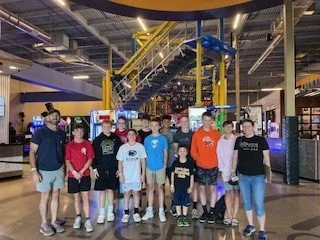 Group of children and teenagers standing together inside an arcade or entertainment center with yellow staircase structure in the background.