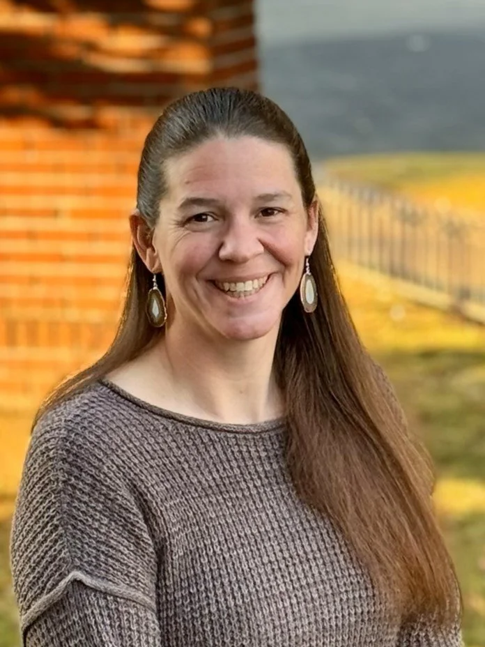 A smiling woman with long brown hair, wearing earrings and a brown textured sweater, standing outdoors near a brick wall and a grassy area in the background.