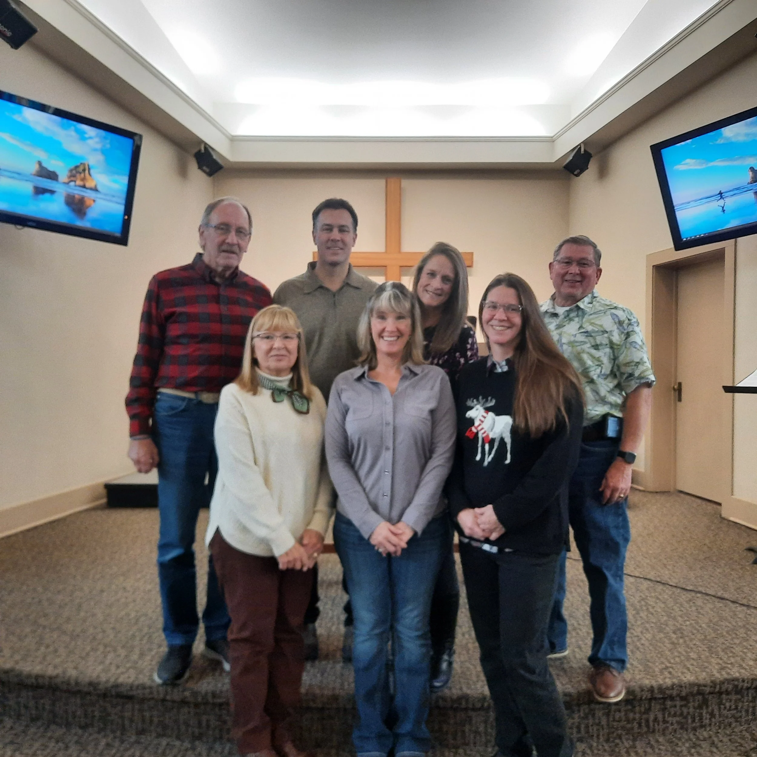 Group of seven people standing inside a church with a wooden cross on the wall behind them.