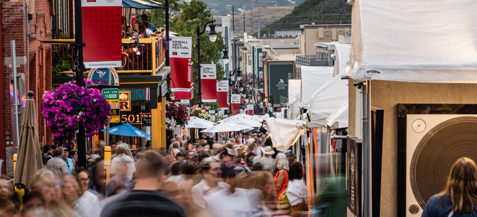 Crowded street scene during a festival with people walking, outdoor tents, flowers, banners, and balcony seating.