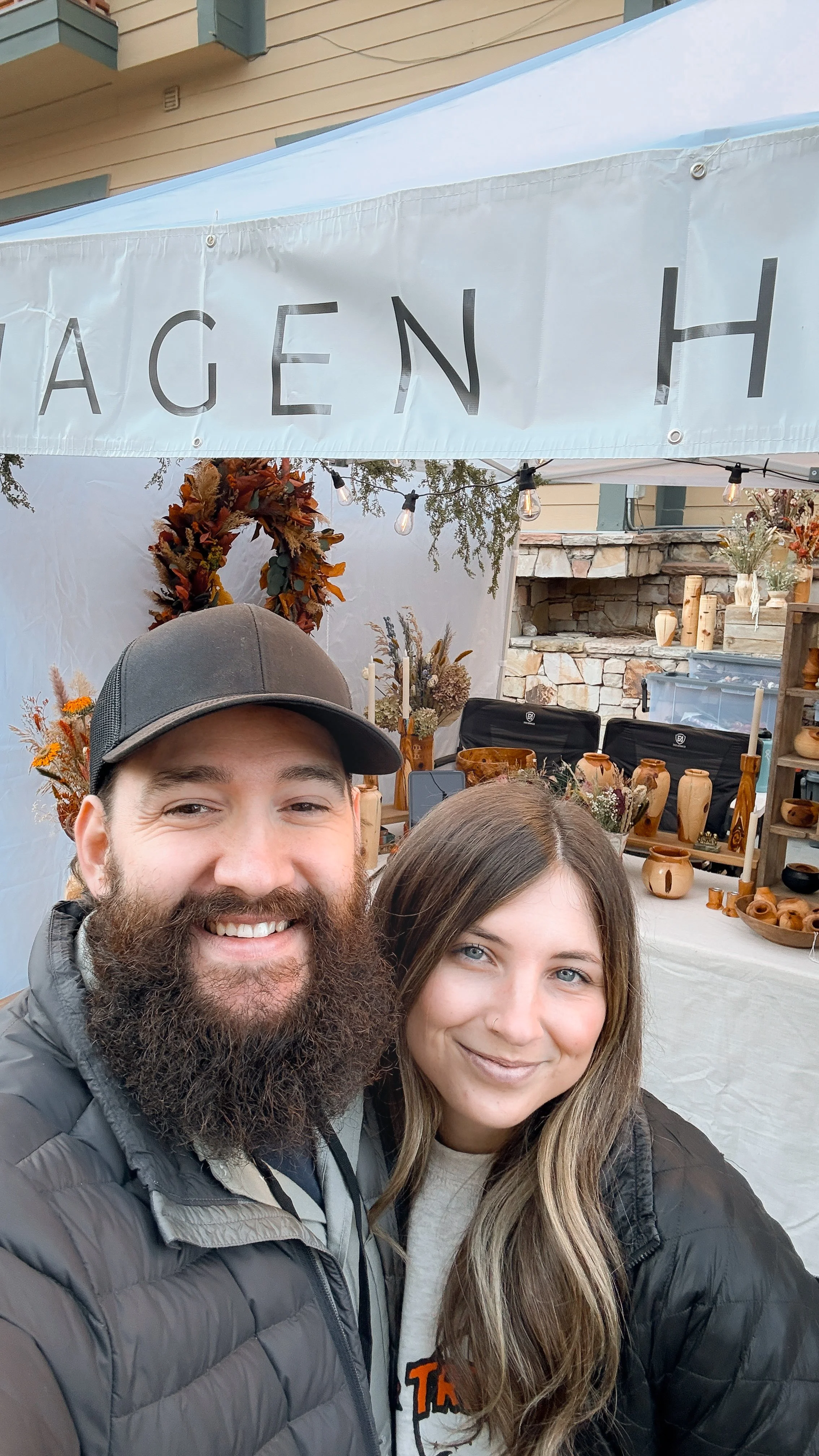 A smiling man with a beard wearing a black cap and jacket, and a woman with long hair and a light-colored shirt, standing in front of a booth with wood vases and dried flower arrangements.