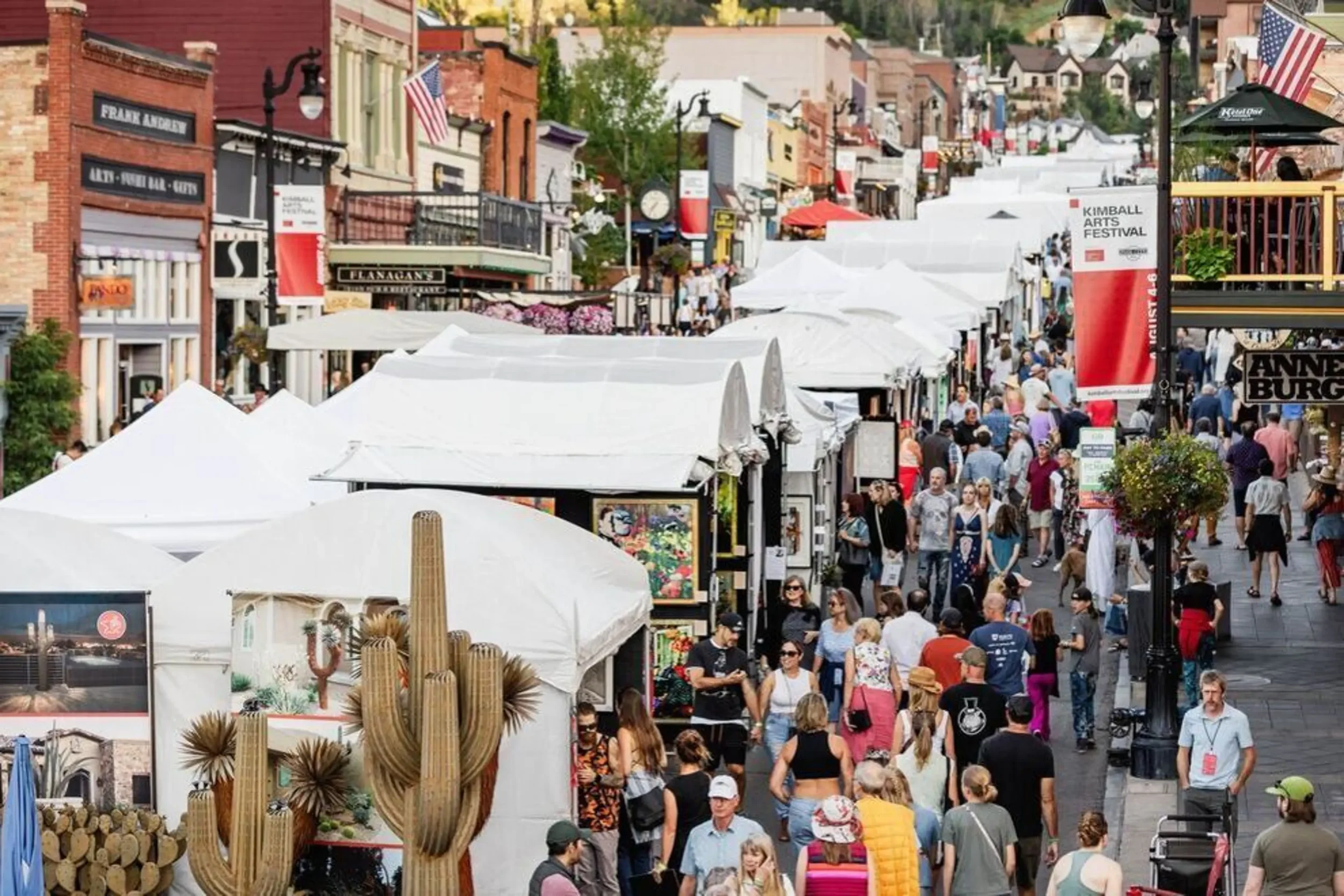 Crowd of people walking along an outdoor festival street lined with white vendor tents and colorful storefronts, with banners and flags overhead.