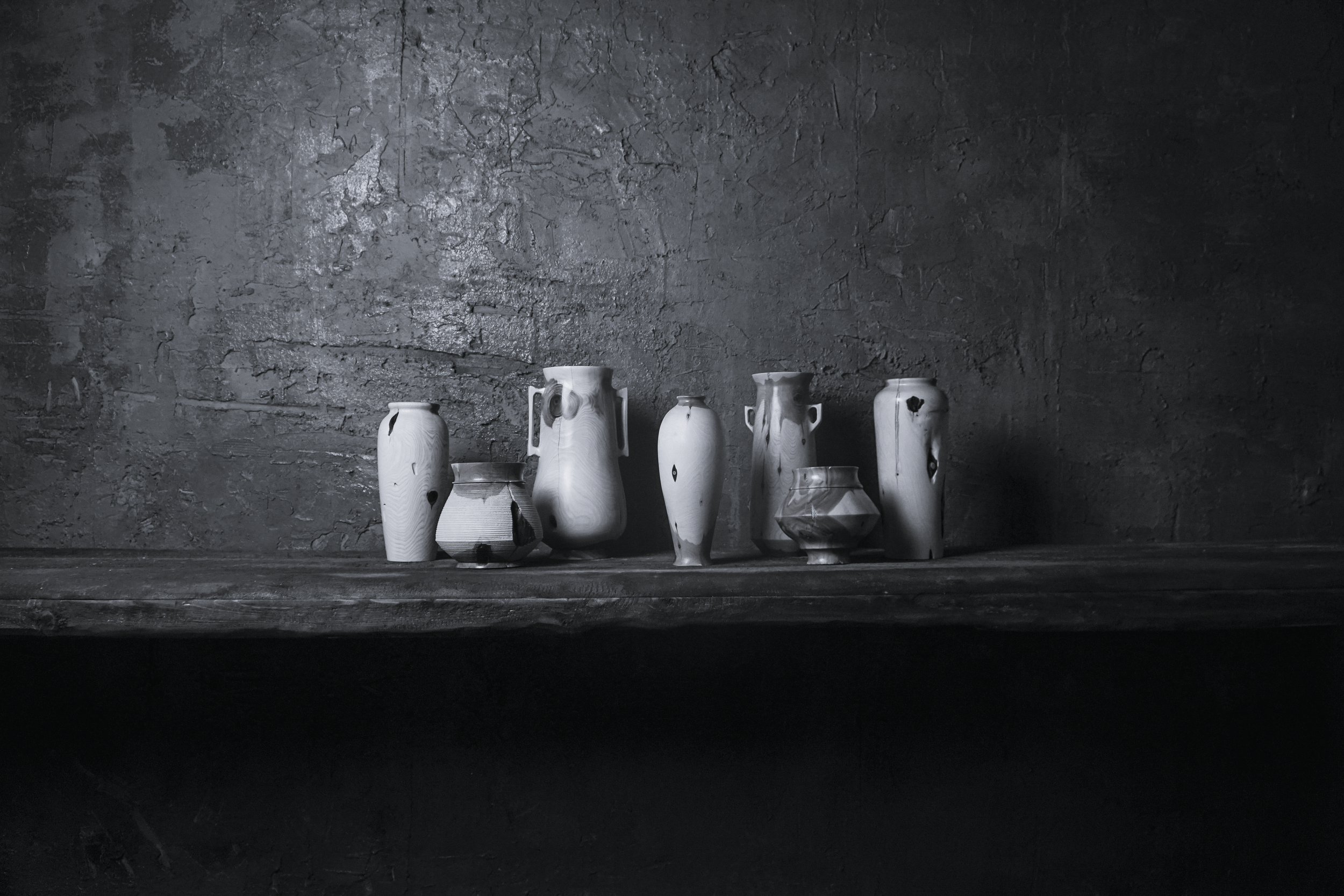 Black and white photo of various ceramic vases and bowls arranged on a wooden shelf against a rough wall.