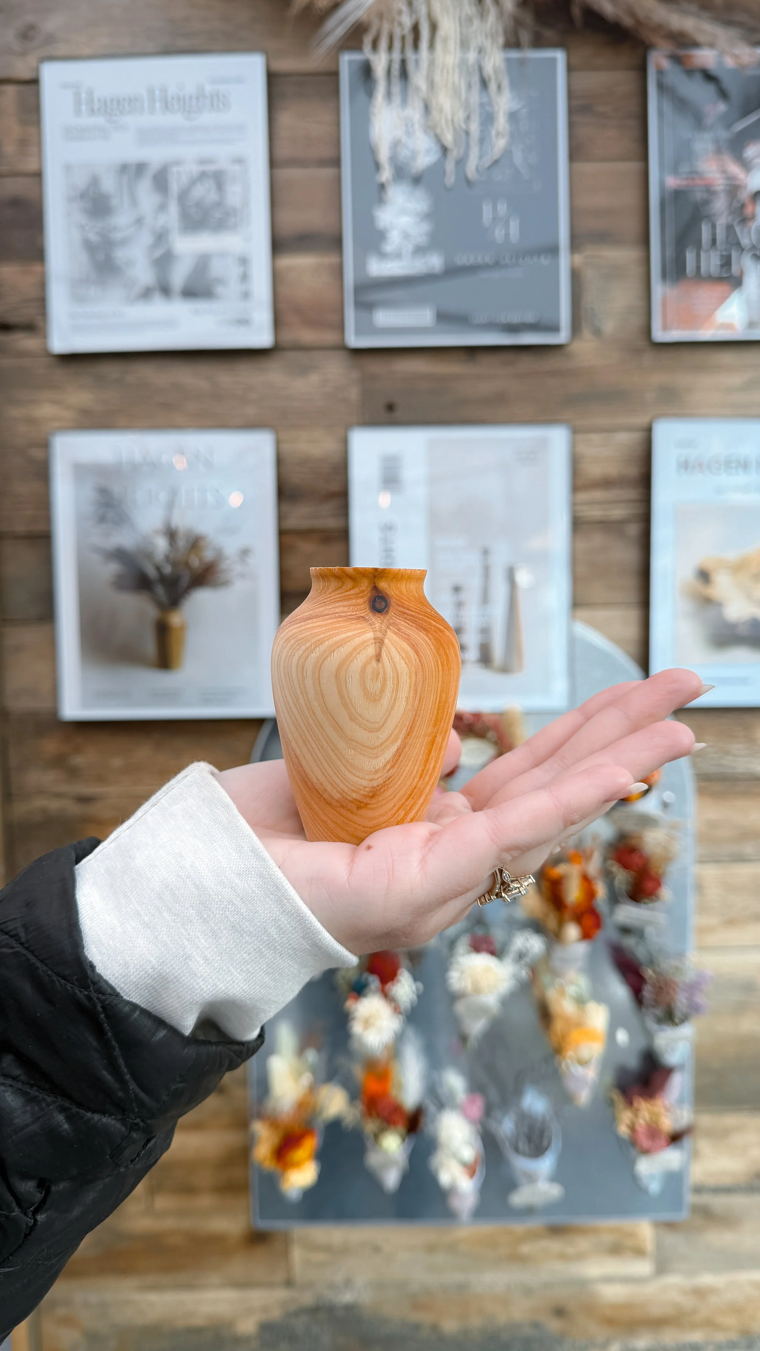 A person holding a small, orange-toned wooden vase inside a store or gallery, with framed posters and a display of dried flowers in the background.