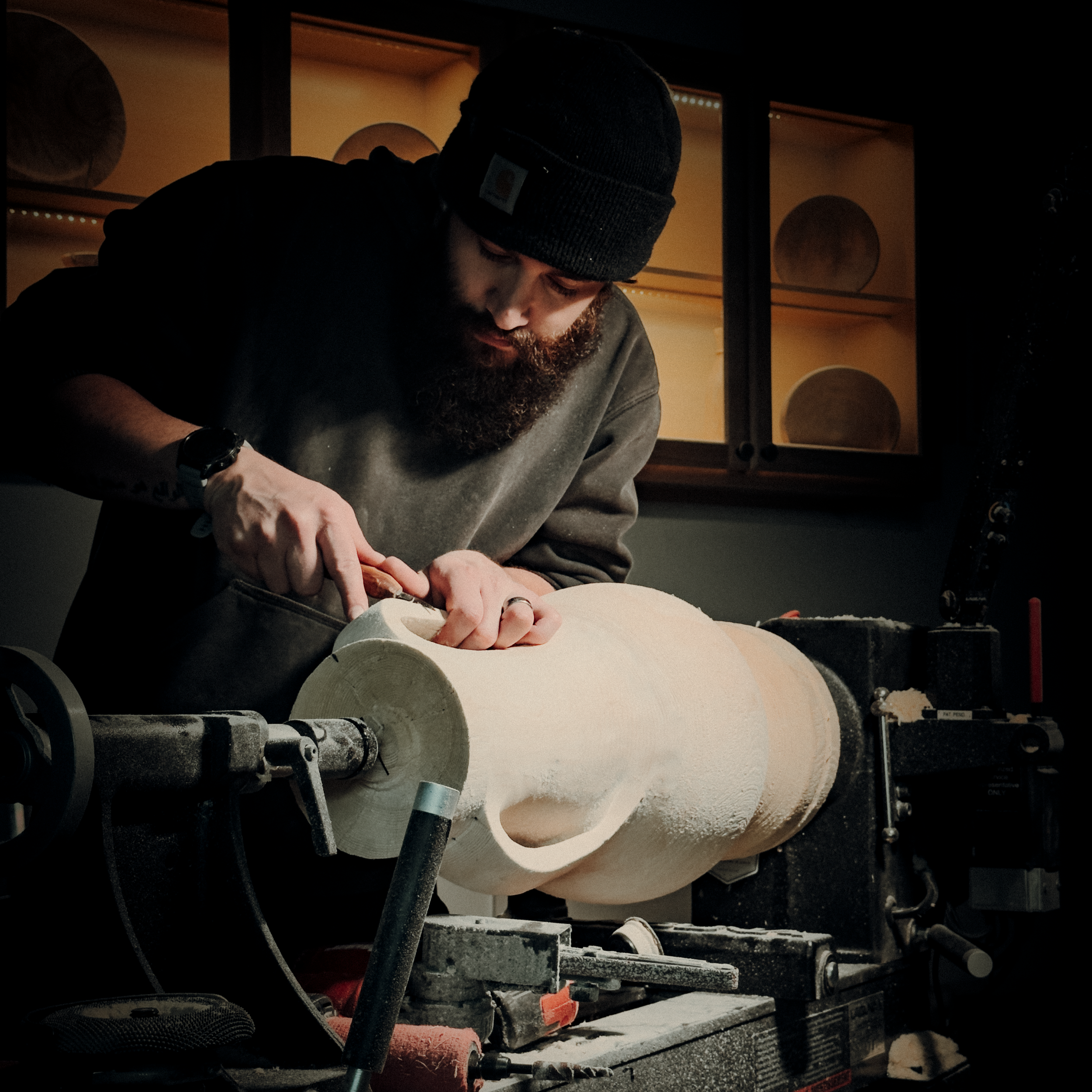 A man with a beard, wearing a black beanie and gray sweatshirt, is using a woodworking tool to carve a large wooden log on a lathe in a workshop.