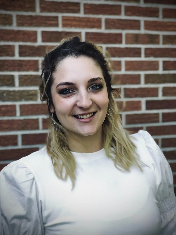 Young woman with blonde hair, dark roots, and blue eyes, smiling in front of a brick wall, wearing a white top with puffed sleeves.