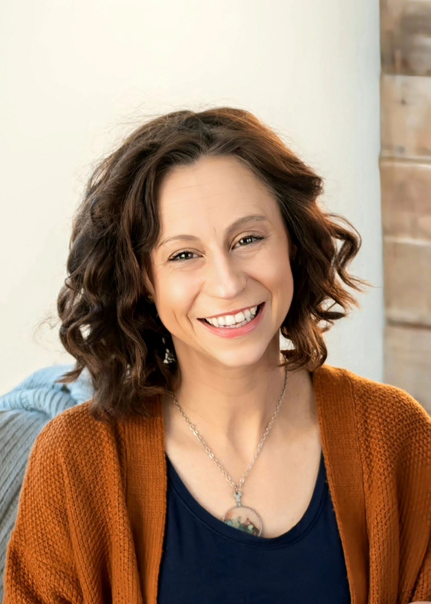 A smiling woman with wavy brown hair wearing a navy blue top, a bronze-colored cardigan, and a pendant necklace.