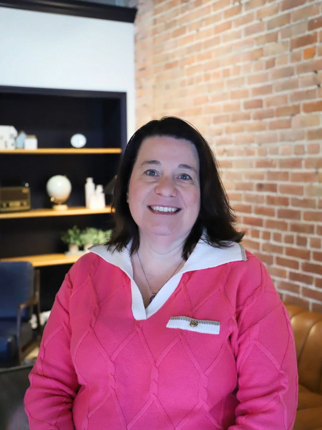 A smiling woman with wavy brown hair wearing a navy blue top, a bronze-colored cardigan, and a pendant necklace.