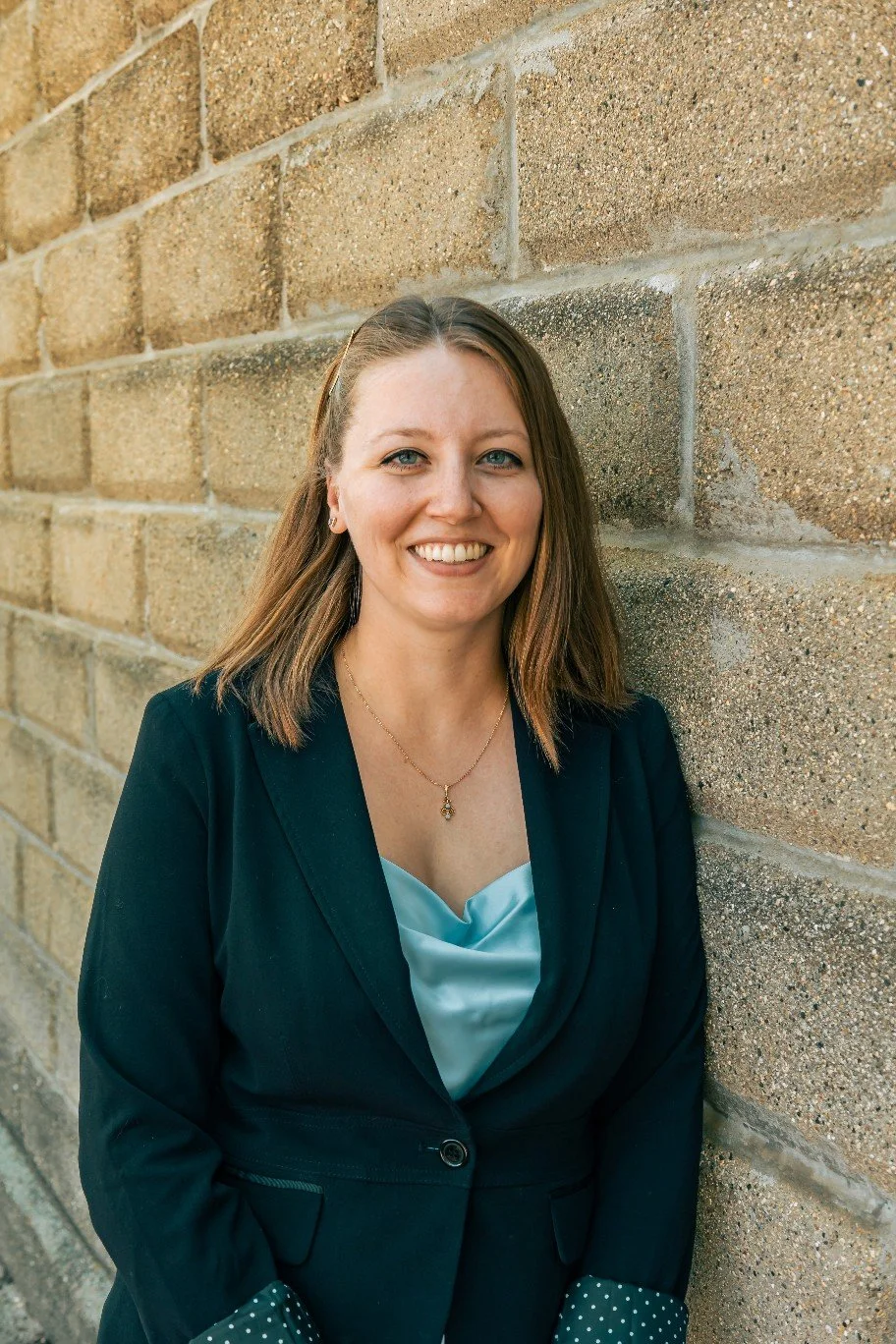 A smiling woman with brown hair in a black blazer and light blue blouse, standing against a brick wall.