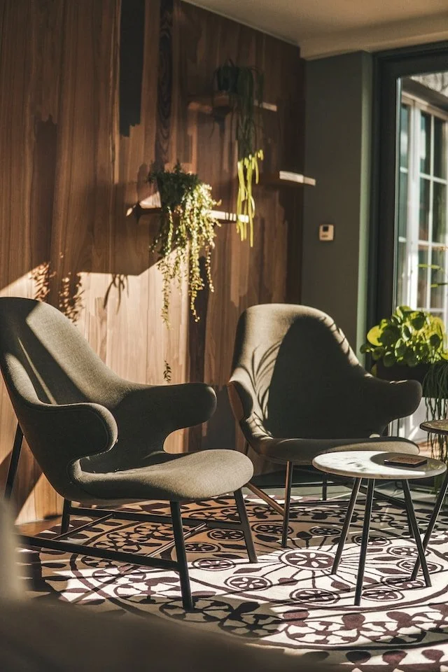 Two dark gray armchairs with wooden legs positioned next to each other in a cozy living room corner, with a small round coffee table with a marble top in front of them, placed on a black and white patterned rug. Behind, there is a wooden accent wall with floating shelves holding plants, and a large window with green curtains allowing sunlight to enter.