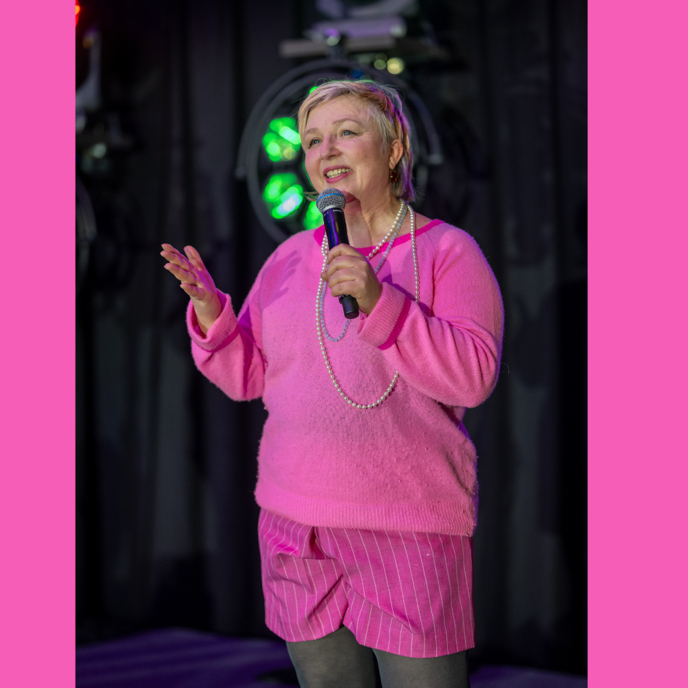 Eine ältere Frau trägt eine pinke Strickjacke, pink-gestreifte Shorts, schwarze Strümpfe und eine Perlenkette, während sie auf einer Bühne mit einem Mikrofon spricht, im Hintergrund bunte Lichteffekte.