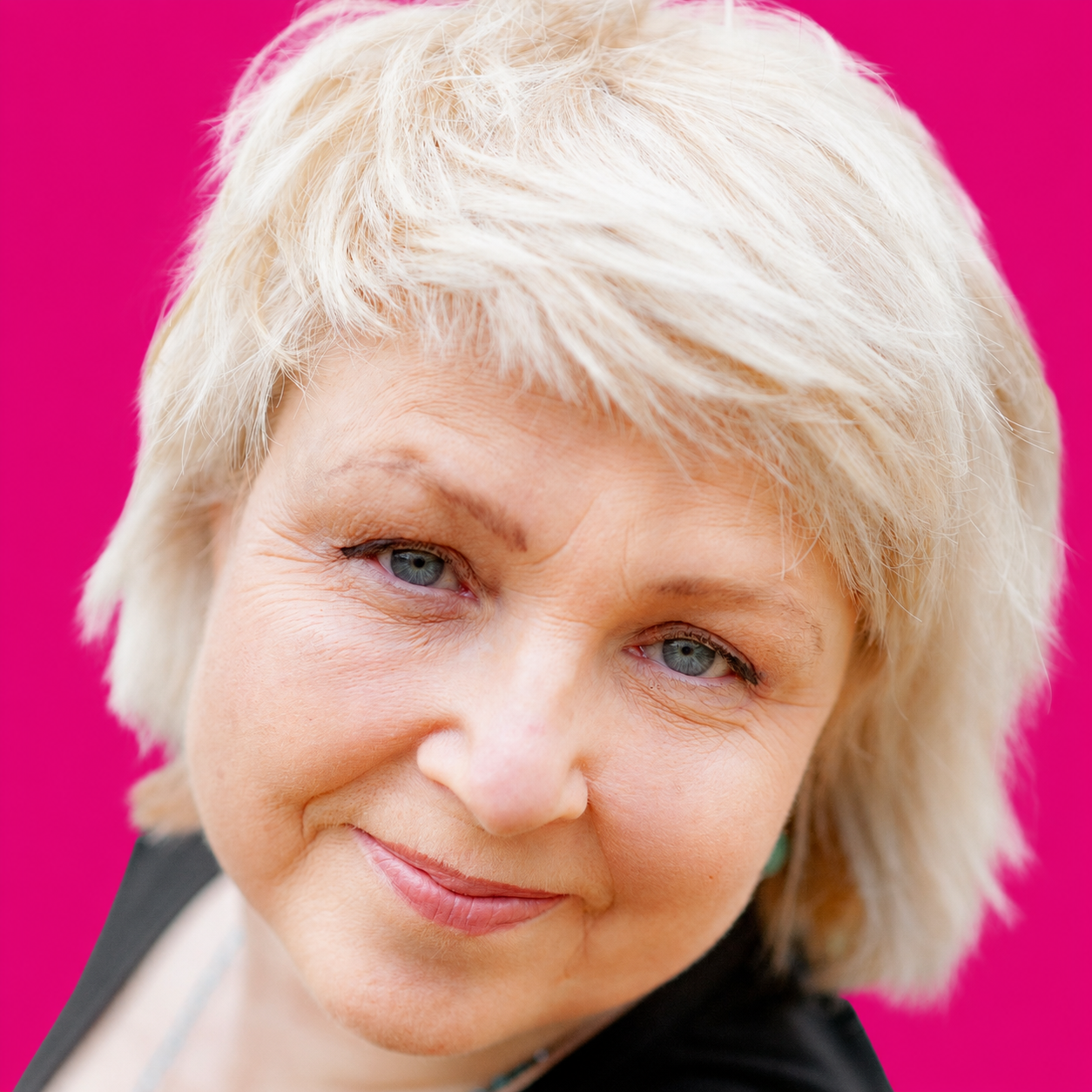 Nahaufnahme eines älteren Frauengesichts mit blonden Haaren und blauen Augen vor pinkem Hintergrund.