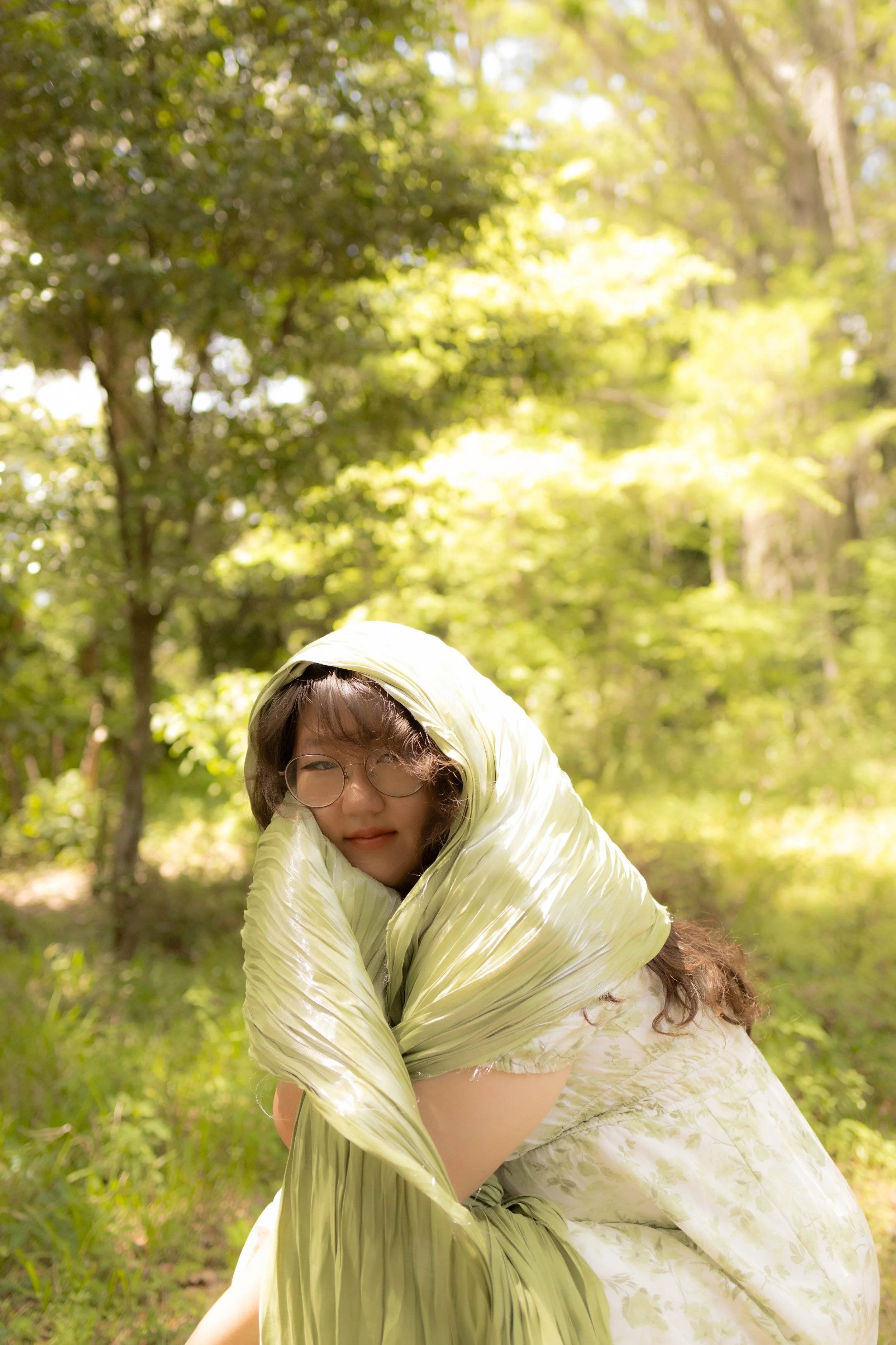 Young woman looking at the camera in the middle of a bright green forest with a shawl over her head.