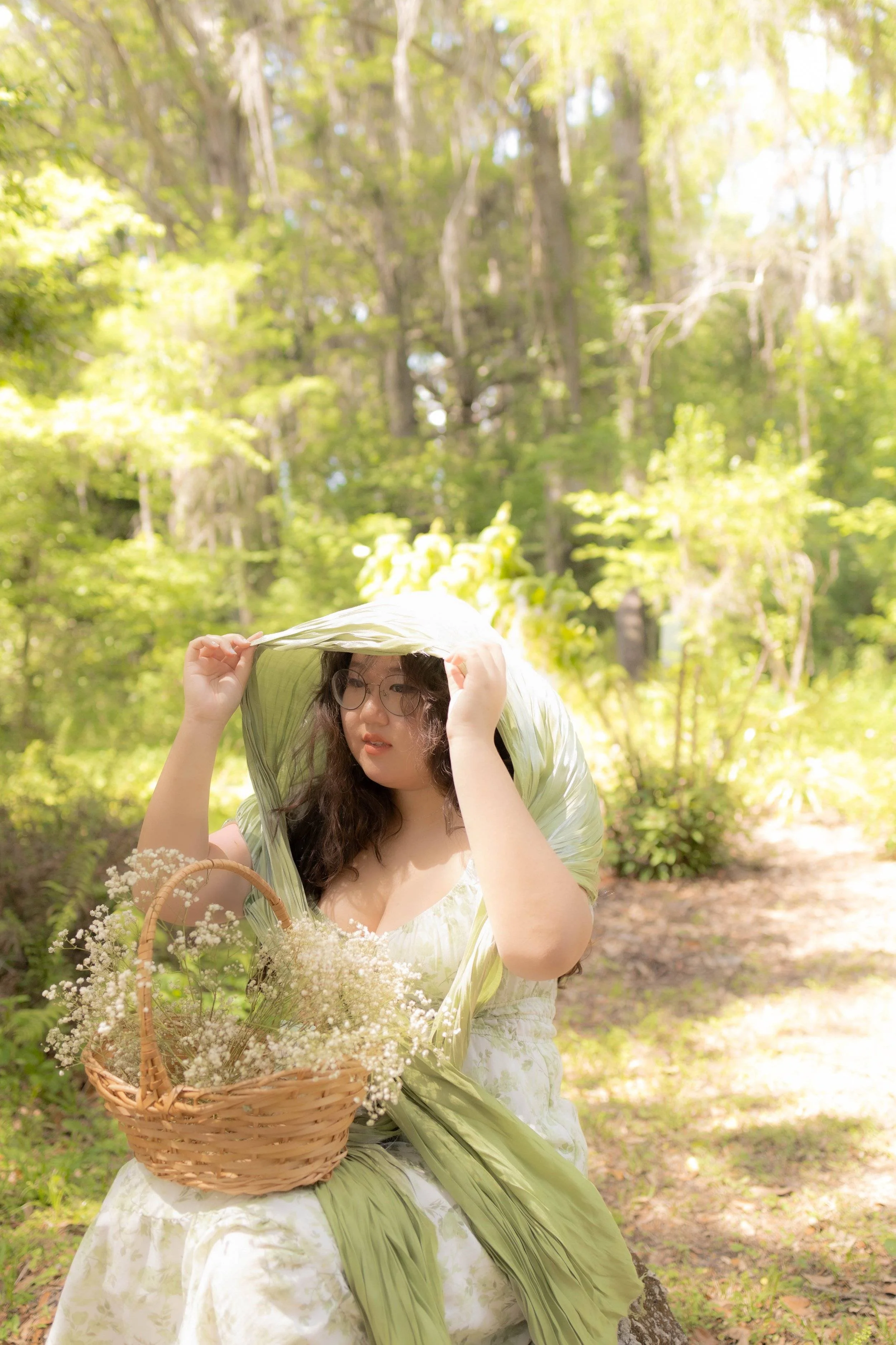 young woman fixing her shawl in a bright green forest with a flower basket on her lap.