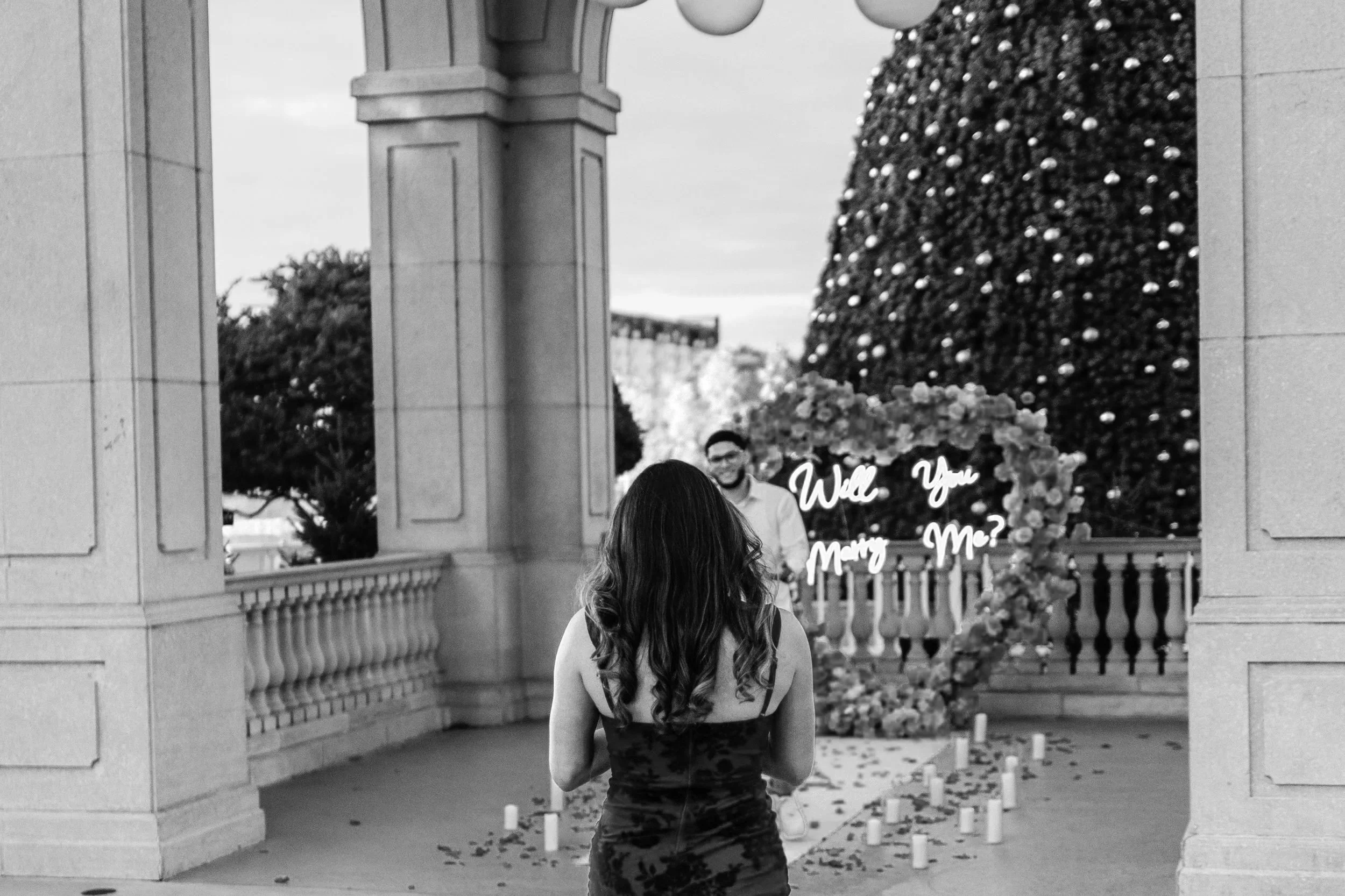 A woman with long hair seen from behind, standing in front of a wedding or engagement photo display with a man in the background. The display includes visible text that reads 'Will You Marry Me?' and decorative flowers, set outdoors with a large Christmas tree decorated with ornaments in the background.