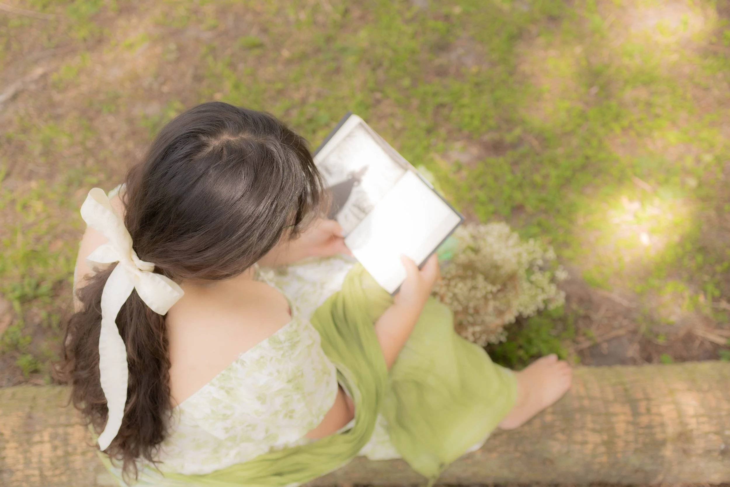young girl barefoot on a log reading a book in a dreamy forest with a cream bow in her hair. 