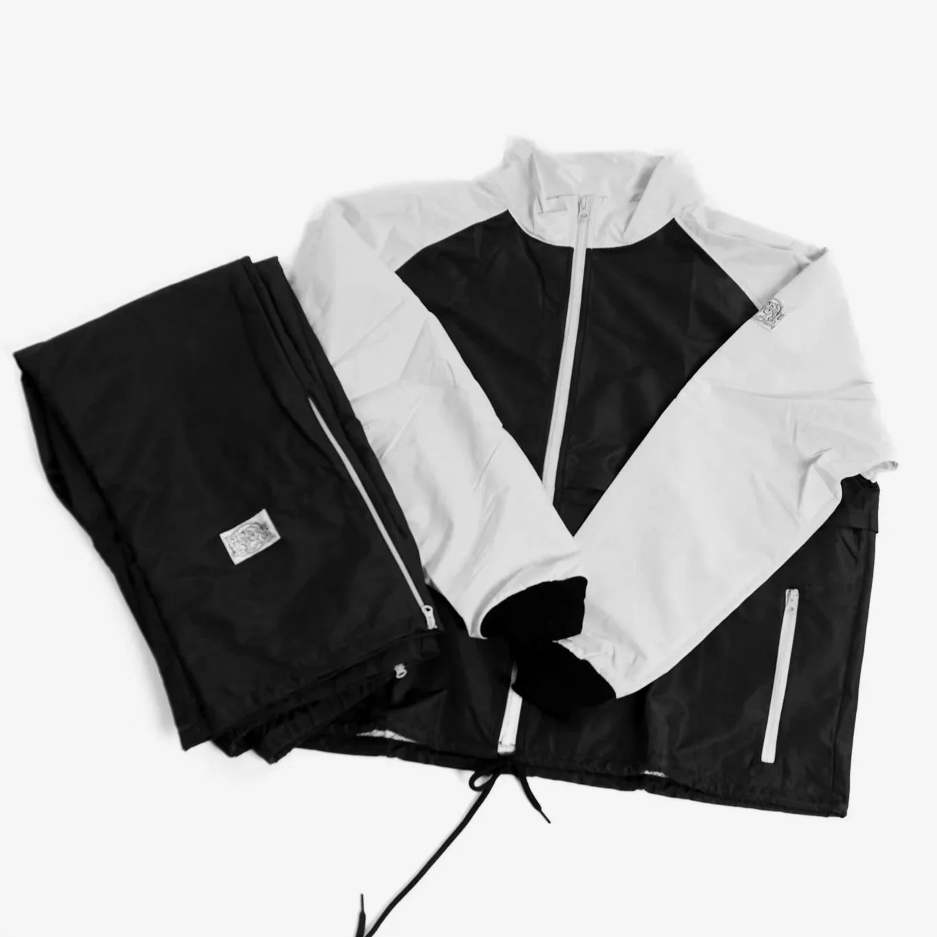 Black and white track jacket with a zip, placed on black pants with a zip pocket, on a white background.