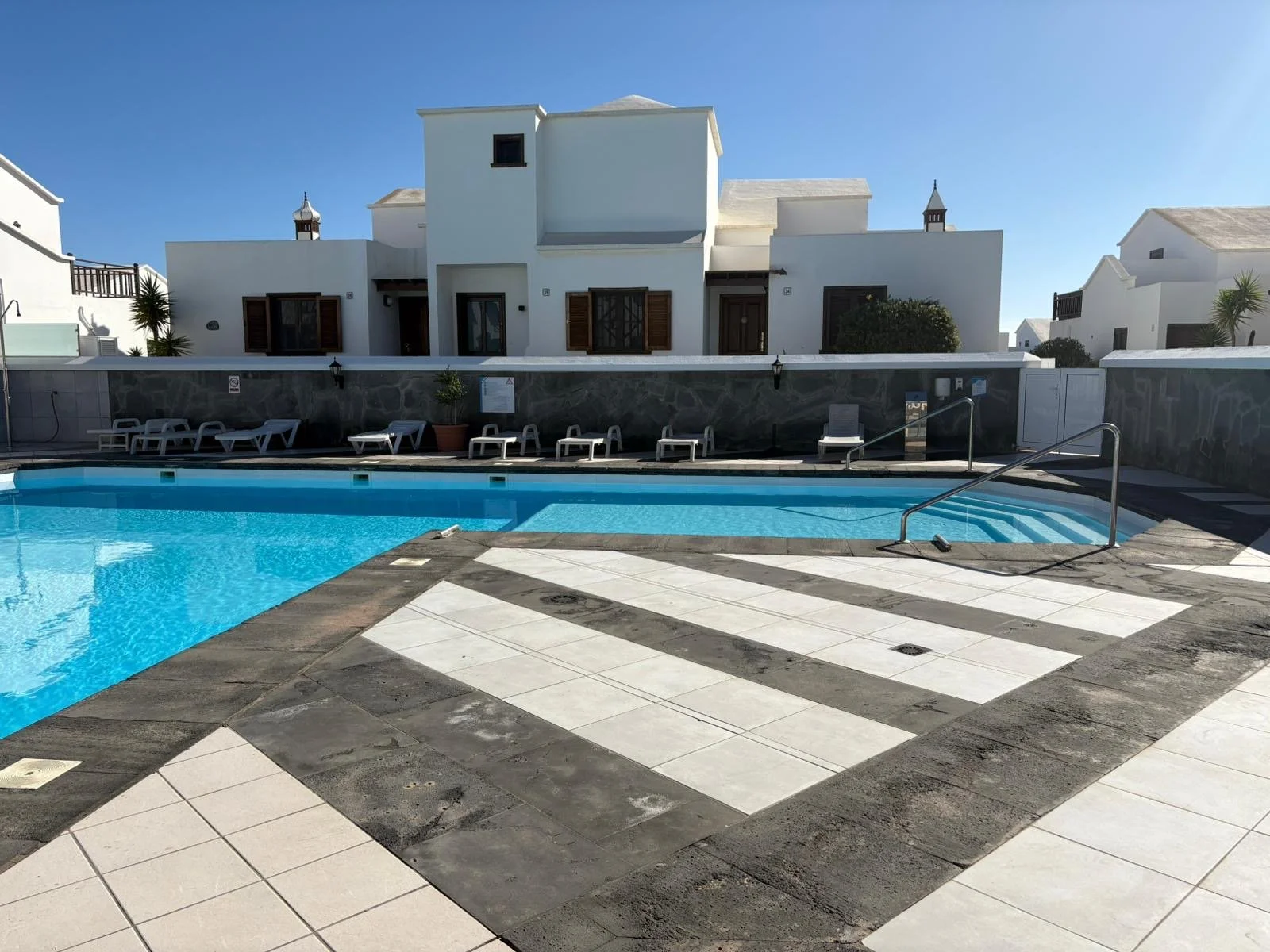 Outdoor swimming pool with deck chairs and a modern white building in the background, under a clear blue sky.