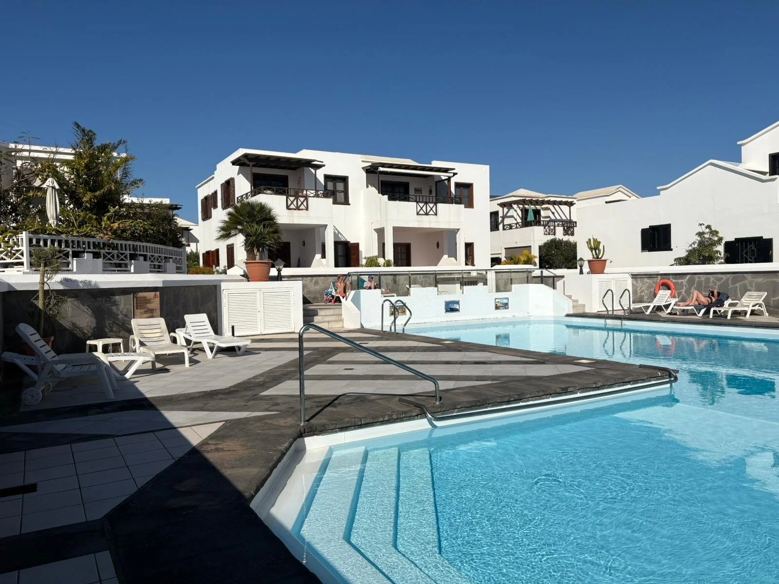 Outdoor swimming pool with surrounding sun loungers, white buildings with balconies, and clear blue sky.