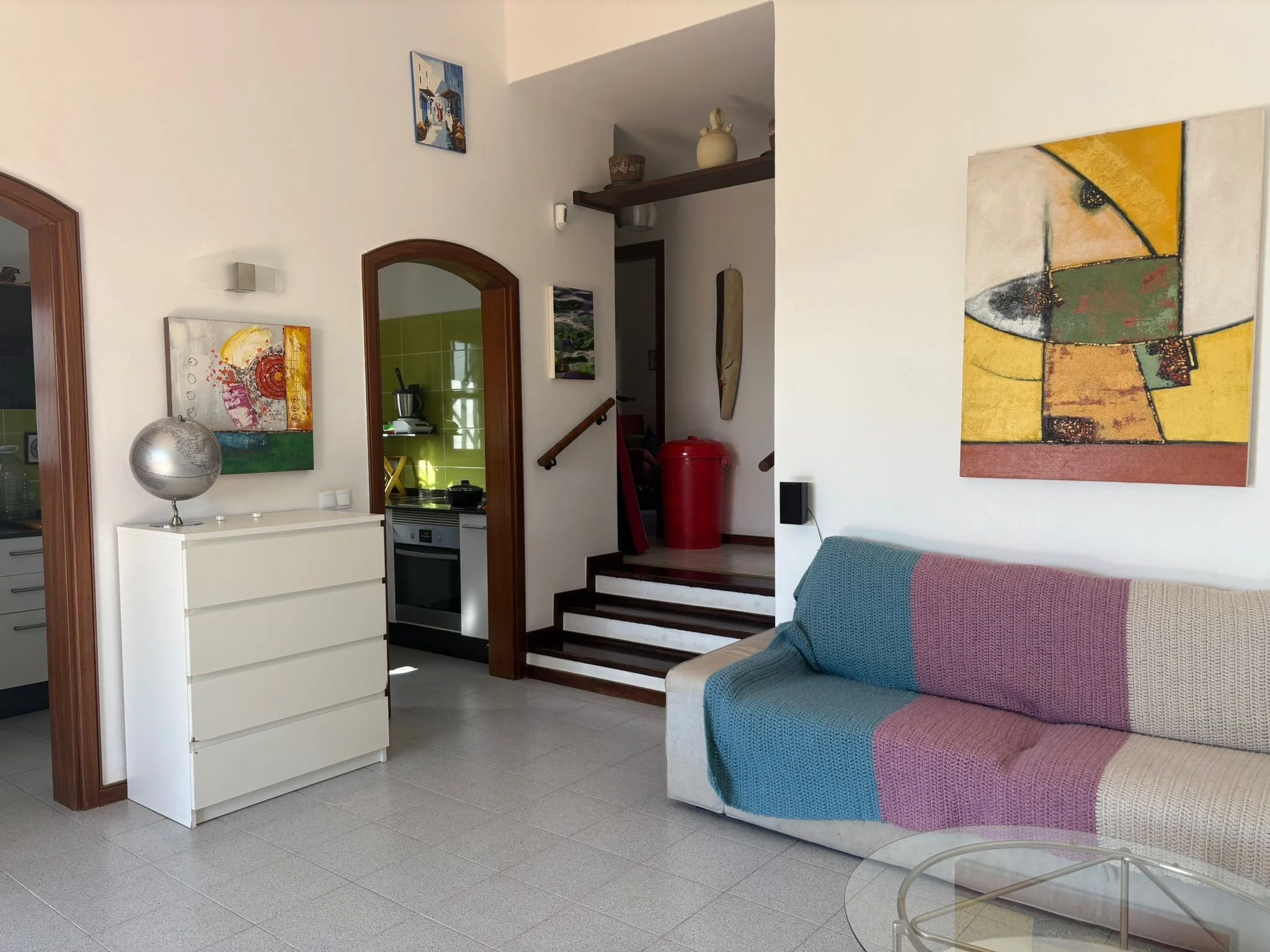 Interior of a modern living room with a white dresser topped with a globe, contemporary abstract paintings on the walls, a beige and pastel-colored sofa with a knitted blanket, a glass coffee table, and an open view into a kitchen area with a red tra