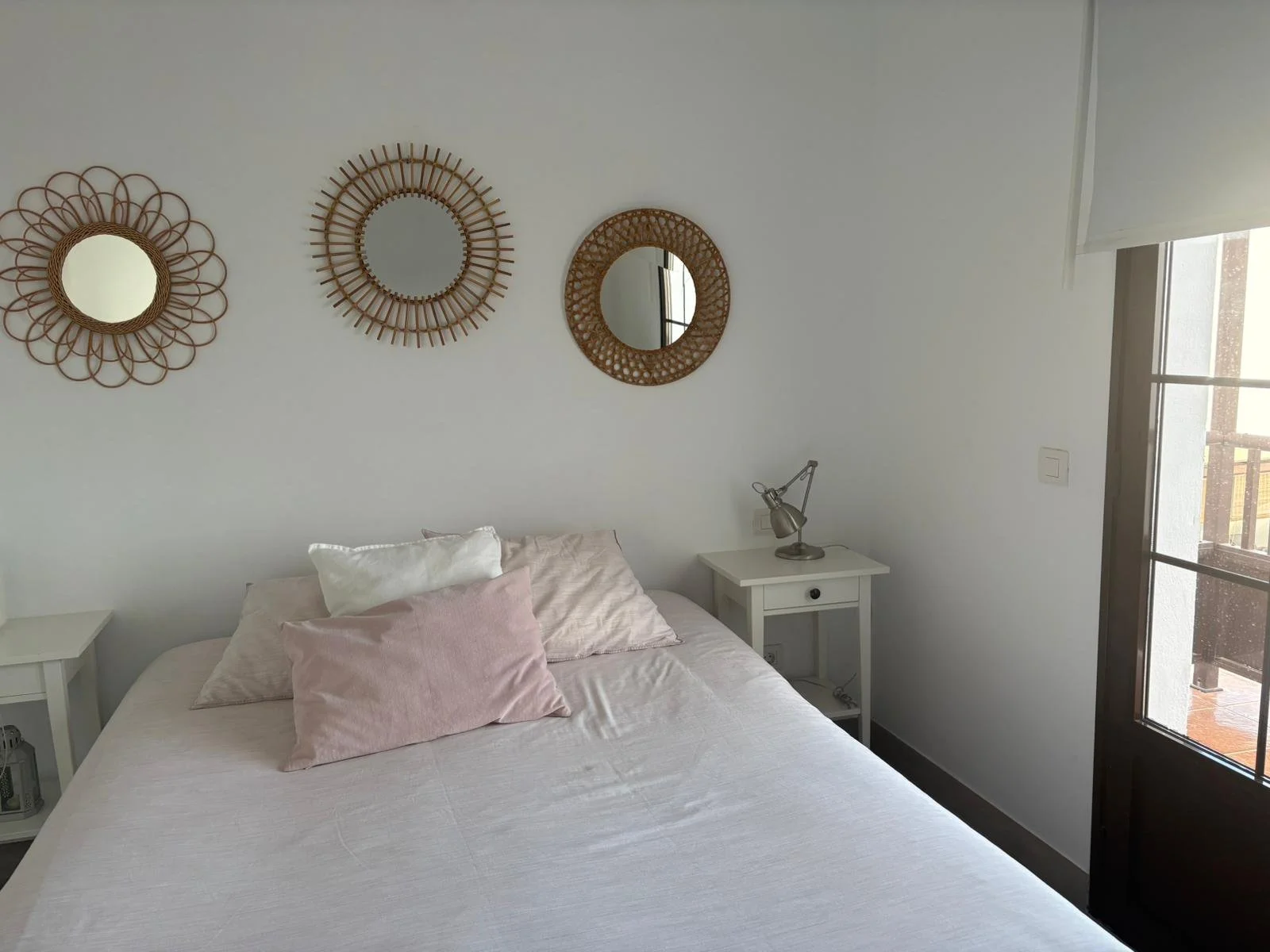 Bedroom with pink bedding, white pillows, three decorative round mirrors on wall, white bedside tables, and a small desk lamp.