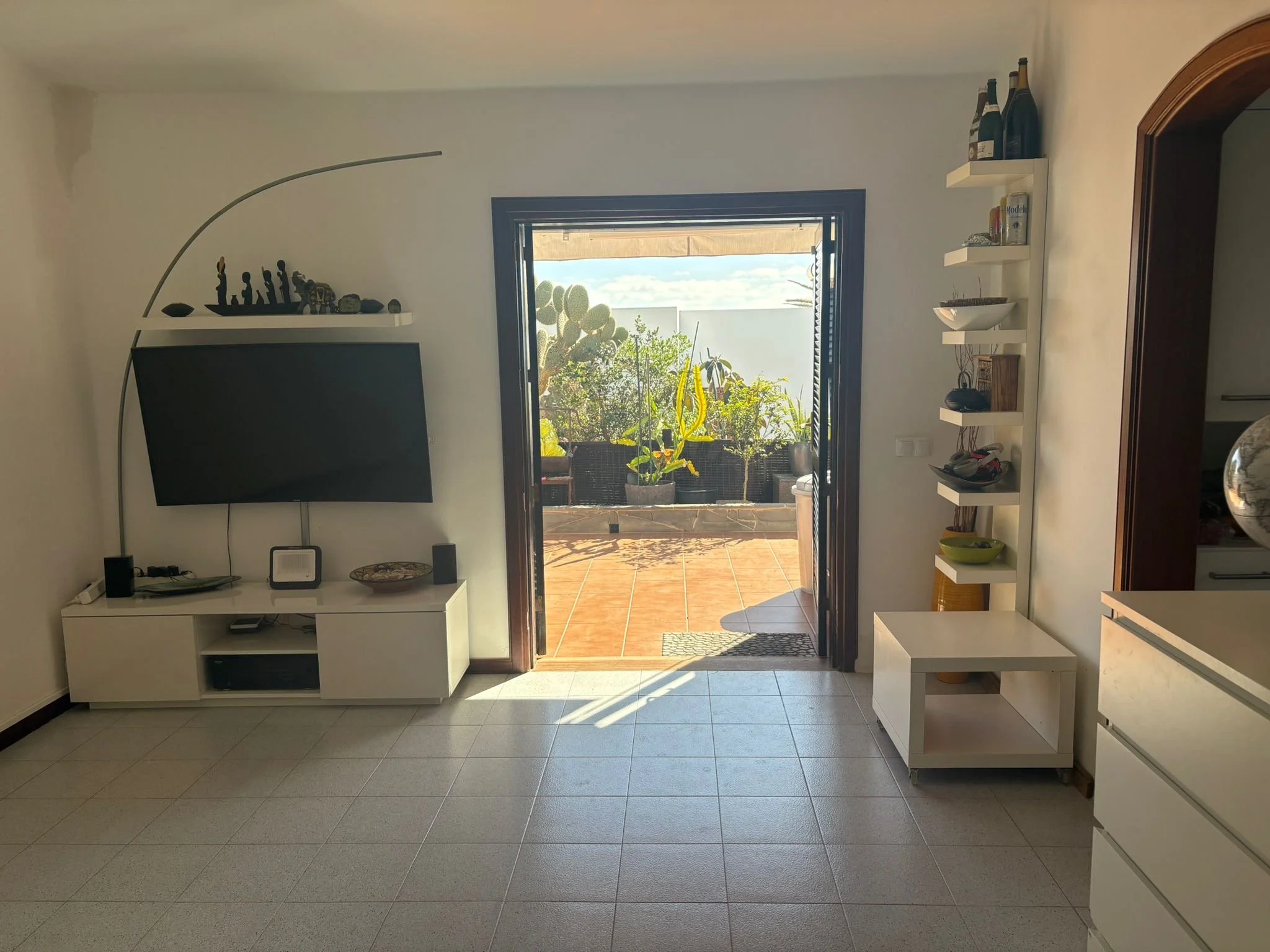 Living room with a flat-screen TV on the wall, white furniture, open doorway leading to a patio with plants. Shelves on the right and a tiled floor.