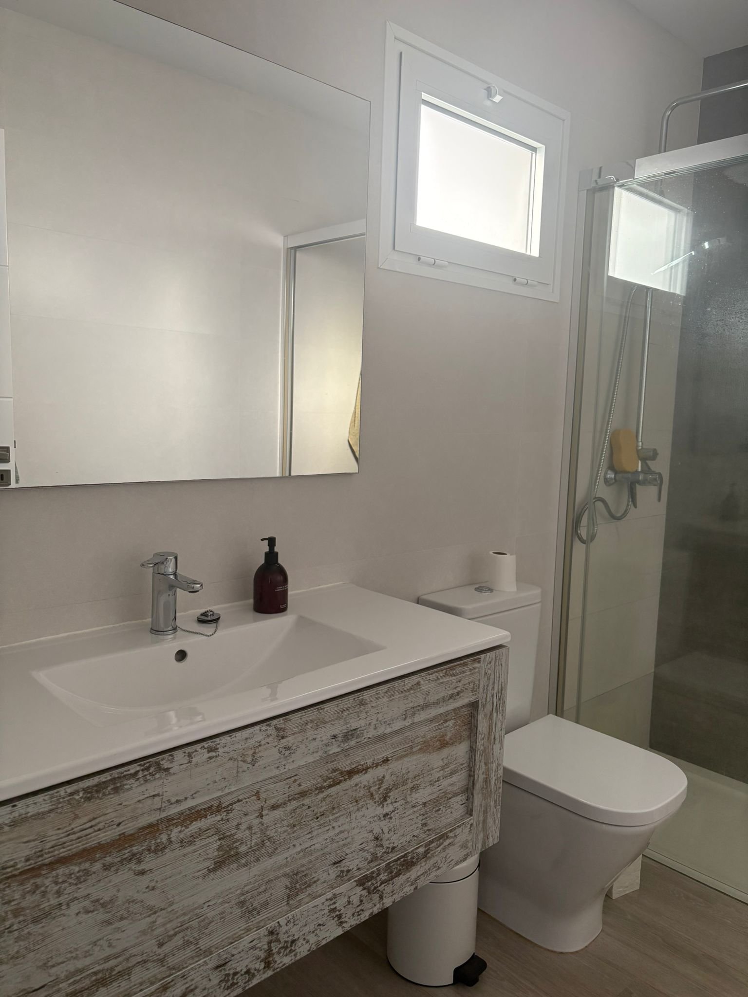 Modern bathroom with a rustic sink, mirror, toilet, small window, and glass shower.