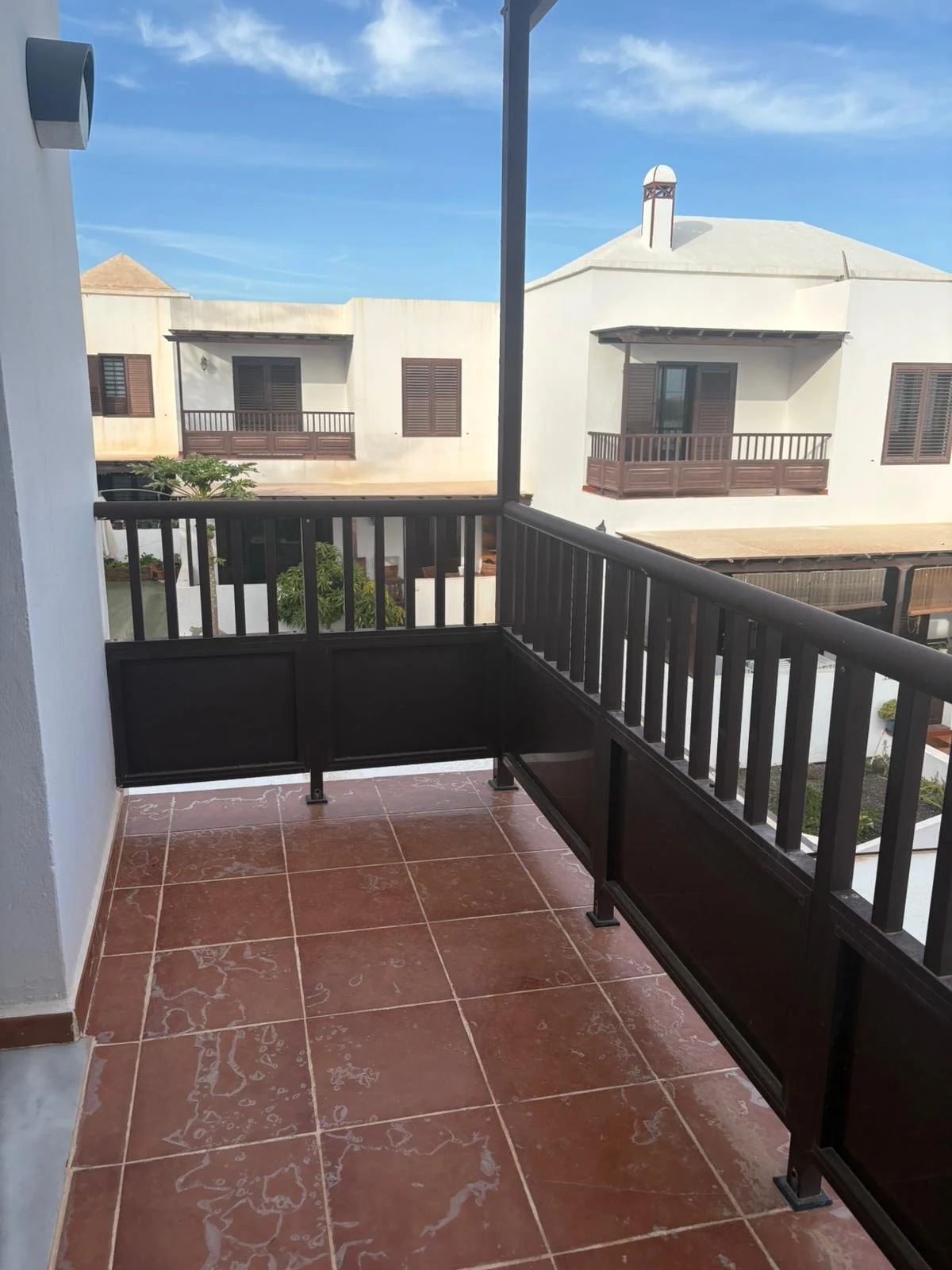 Outdoor balcony with brown tiles and a wooden railing, facing neighboring houses with white walls and dark shutters under a blue sky with wispy clouds.