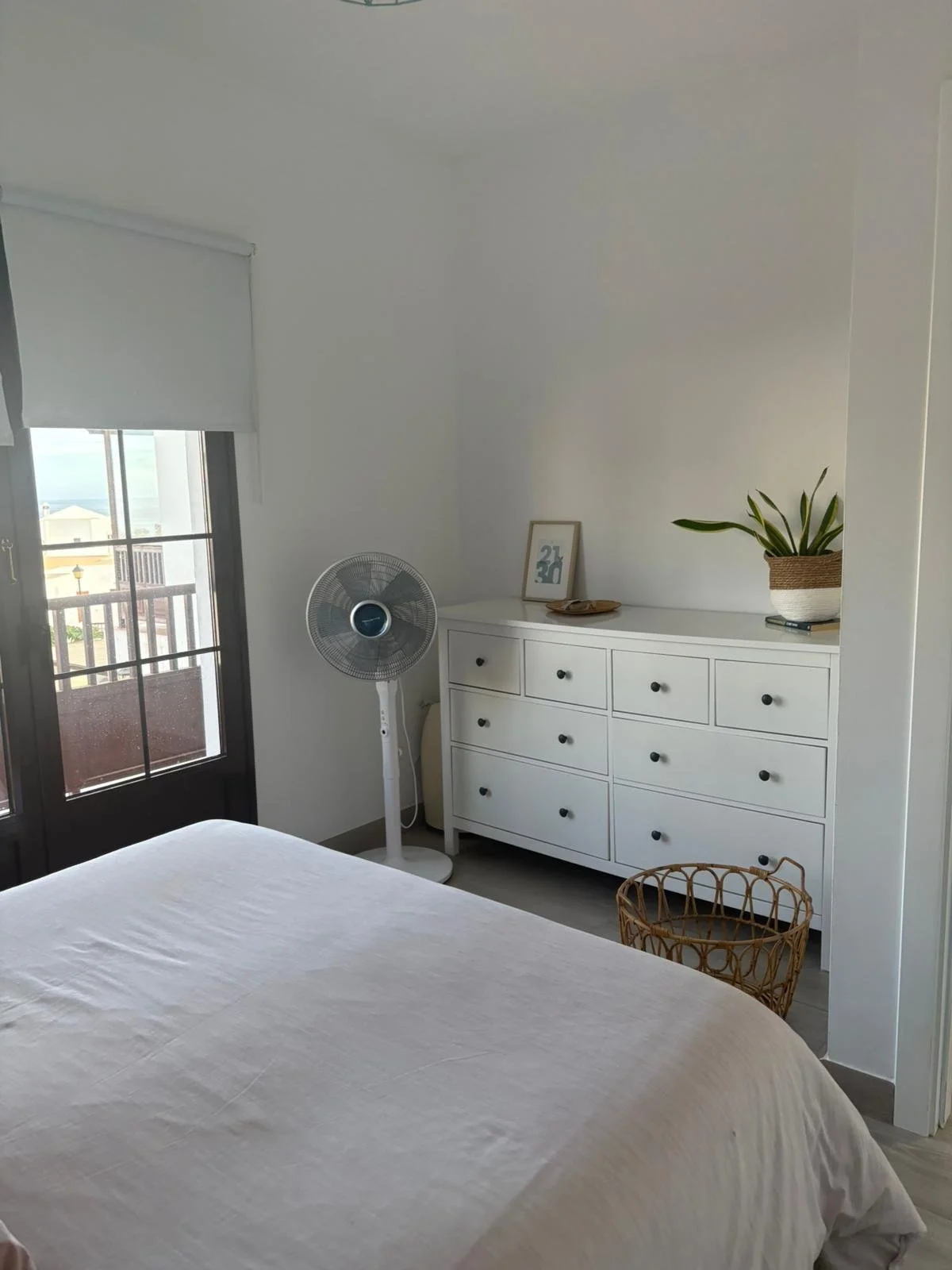 Minimalist bedroom with a white dresser, potted plant, framed artwork, standing fan, woven basket, bed with gray bedding, and a window with closed blinds.