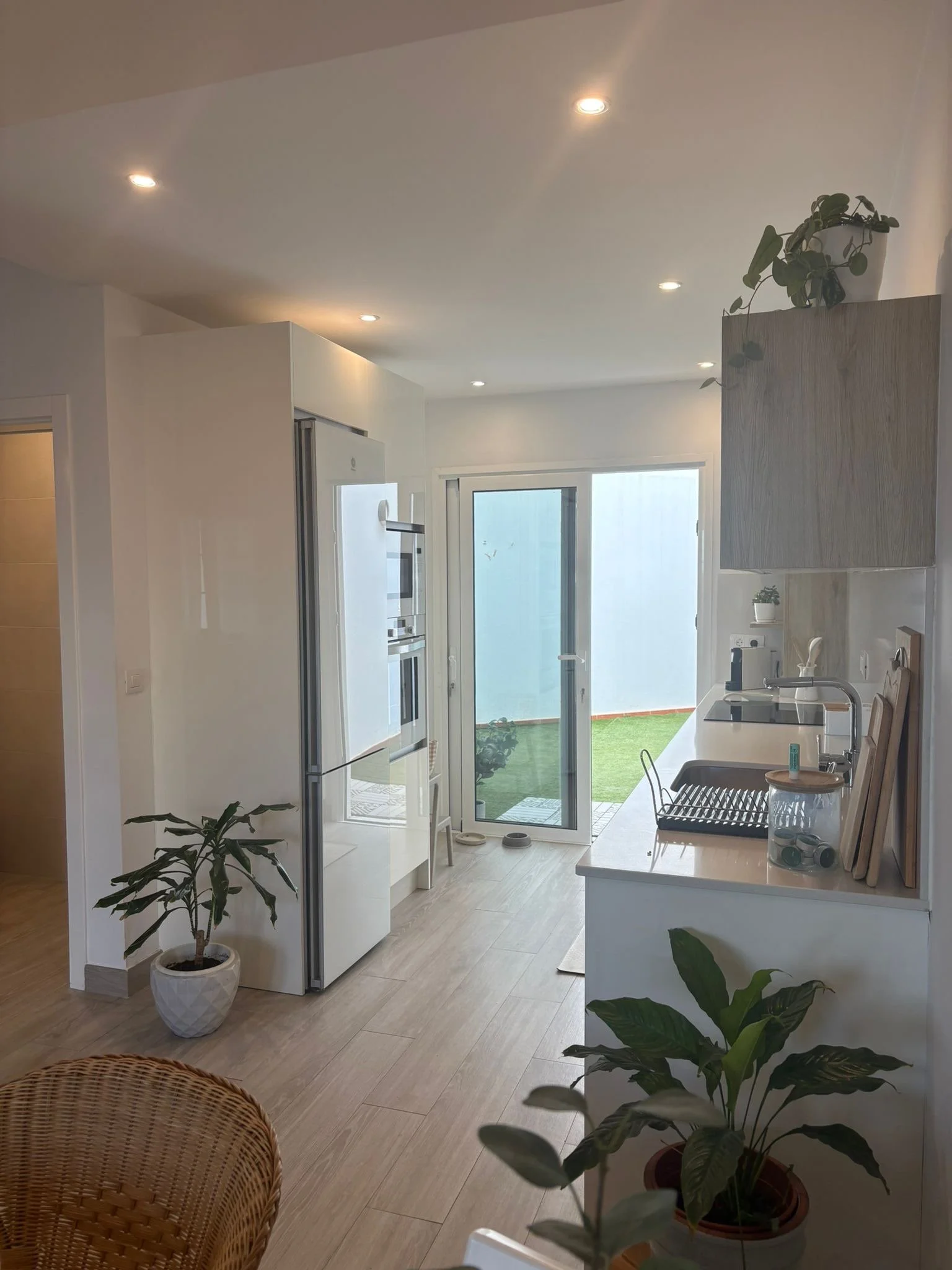 Modern kitchen with light wood flooring, white cabinets, potted plants, and natural light from large sliding glass doors leading to a patio with green grass.