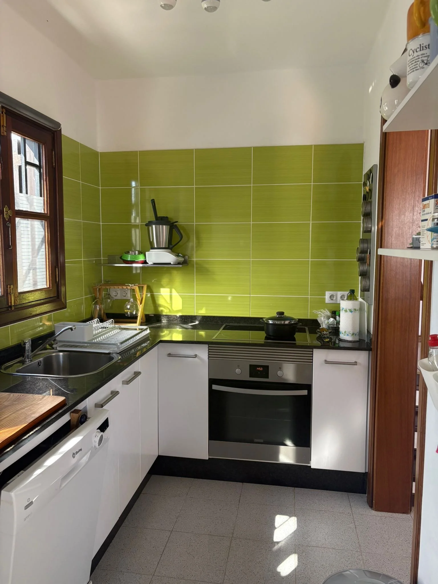 Modern kitchen with white cabinets, green tiled backsplash, stainless steel appliances, and a small window. Features include a sink, oven, induction cooktop, and various kitchen utensils.