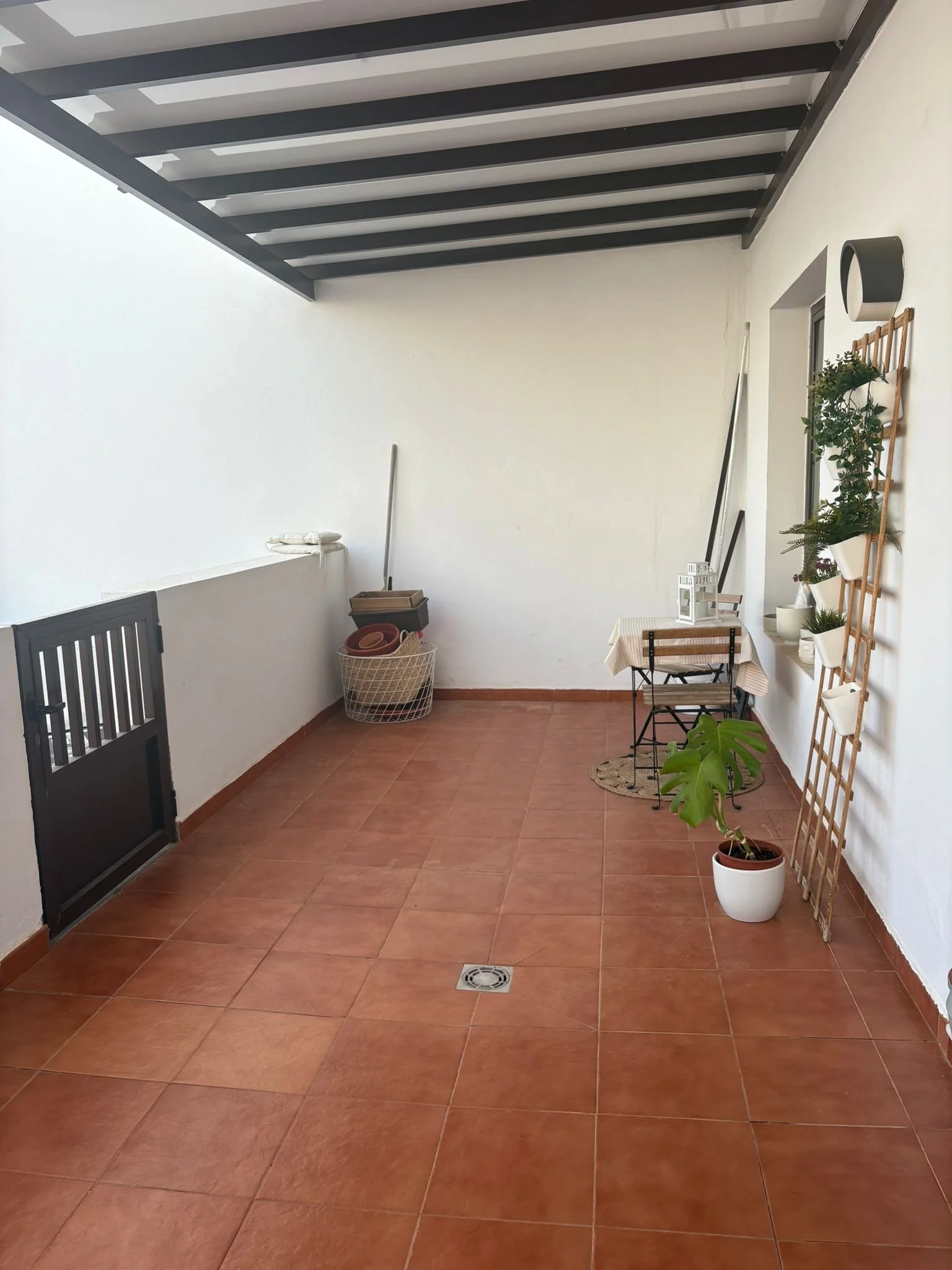 Outdoor terrace with terracotta floor tiles, black wooden beams overhead, a small table with a chair, potted plants, a lattice with greenery, and a broom. There are also storage baskets and various decor elements.