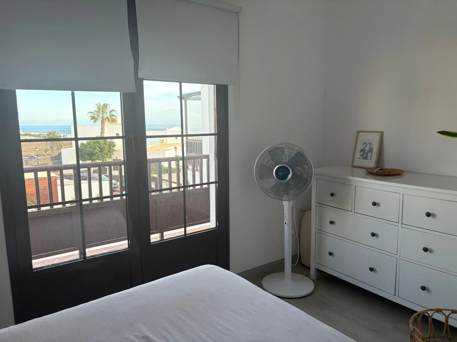 Bedroom with a view of the ocean through glass doors, featuring a white dresser with drawers, a standing fan, framed artwork on the dresser, and a neatly made bed with a white sheet.