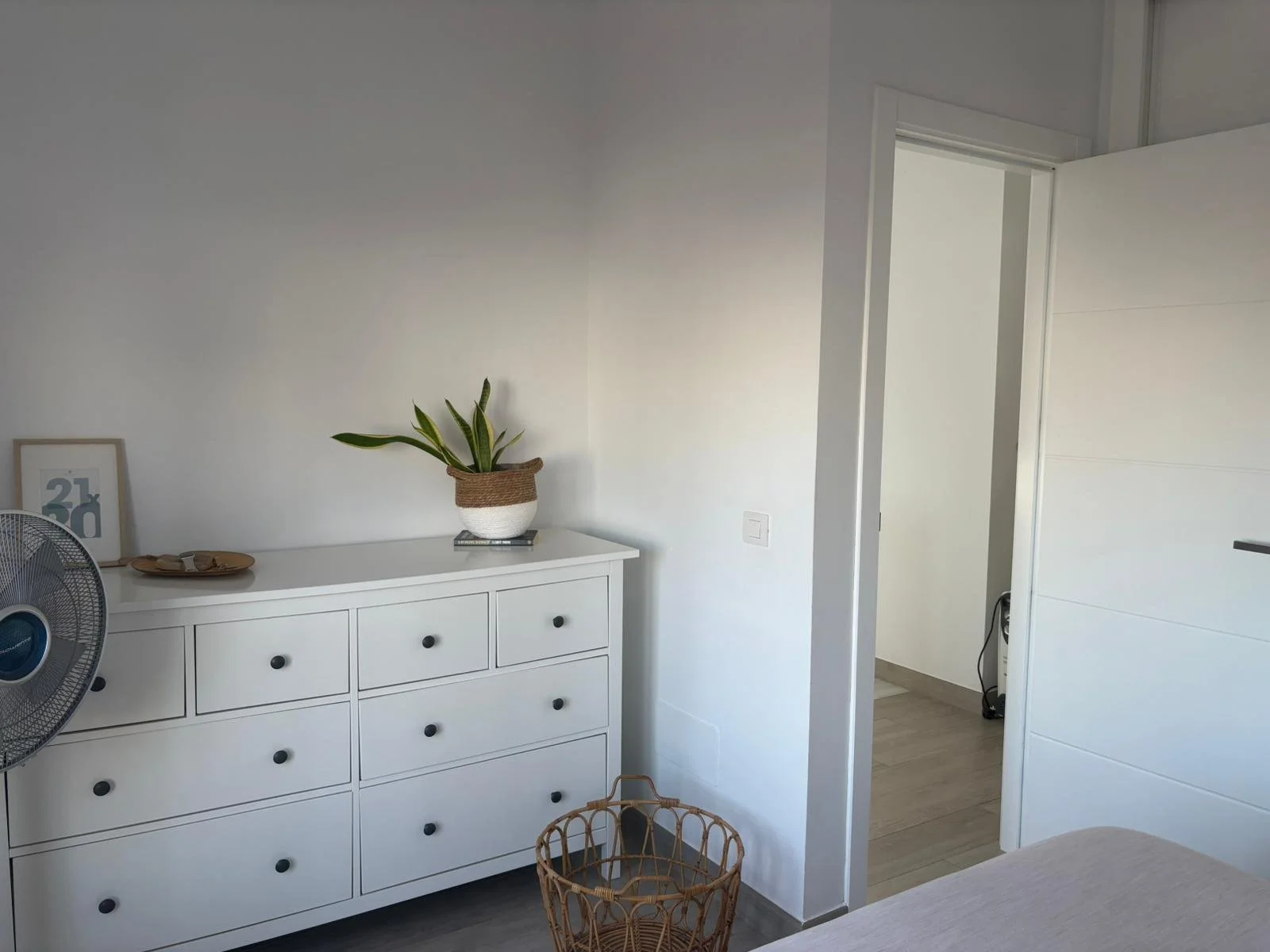 Minimalist bedroom with a white dresser, potted plant, standing fan, and open door.