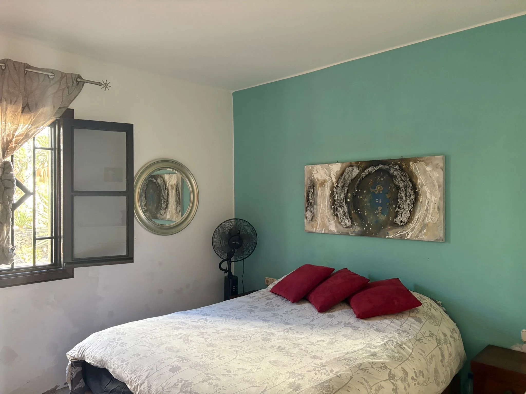 Bedroom with turquoise accent wall, bed with patterned duvet and red pillows, window with sheer curtains, round mirror, standing fan, and abstract wall art.