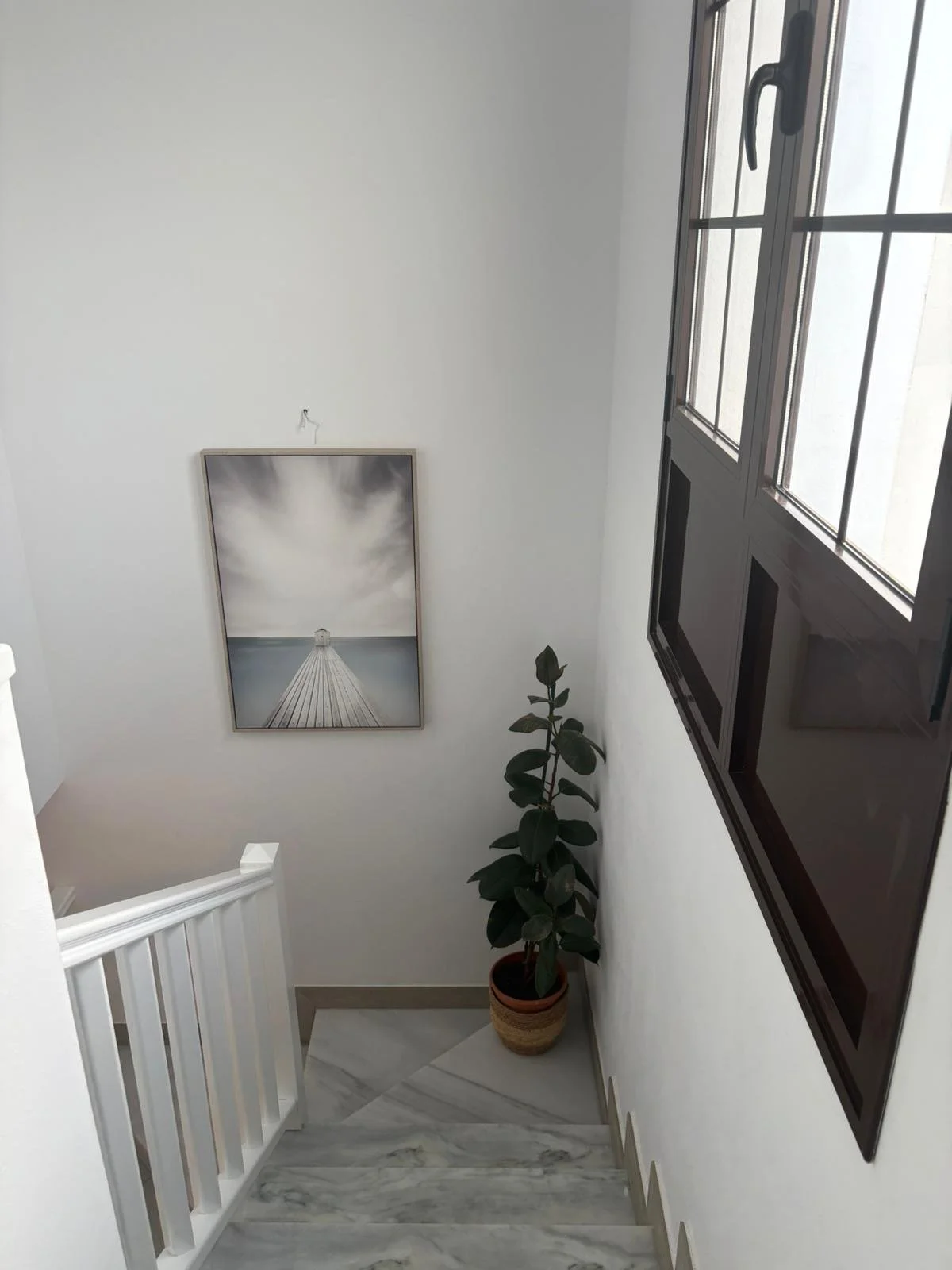 Indoor staircase with a potted plant, a large window, a framed photo of a pier, and white walls.