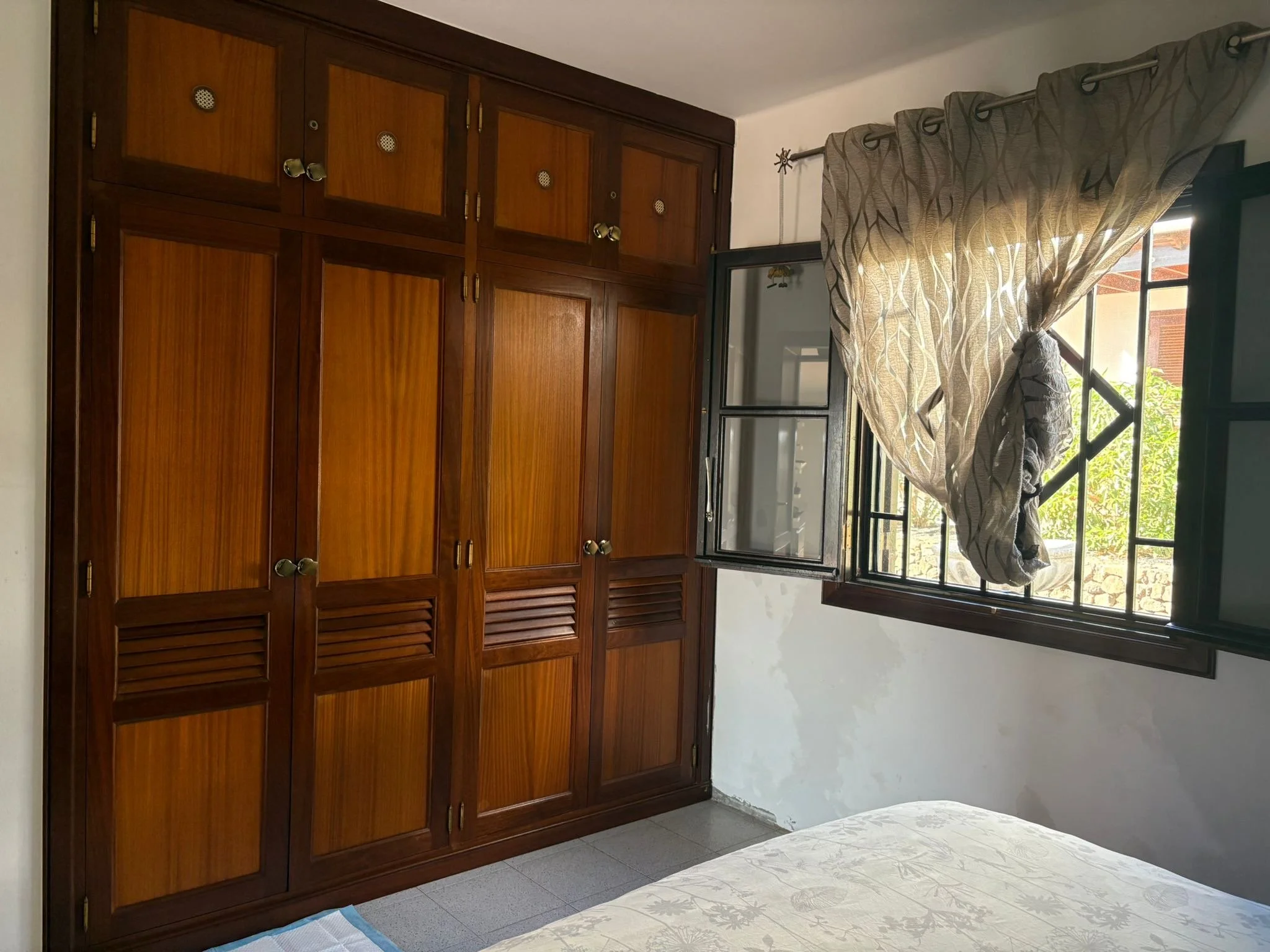 Bedroom with large wooden wardrobe, open window with patterned curtain tied back, and partial view of a bed.