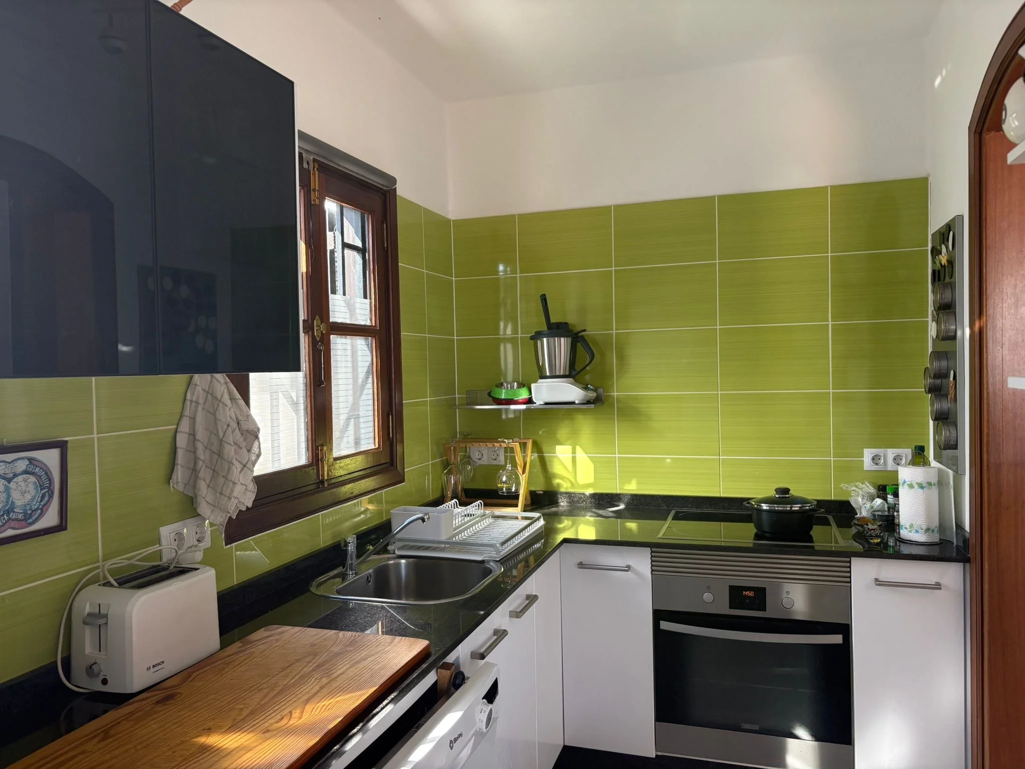 Modern kitchen with green tile backsplash, white cabinets, wooden window, and countertop appliances. Features include a toaster, food processor, sink with dish rack, stovetop with pot, and wall-mounted utensil holder.