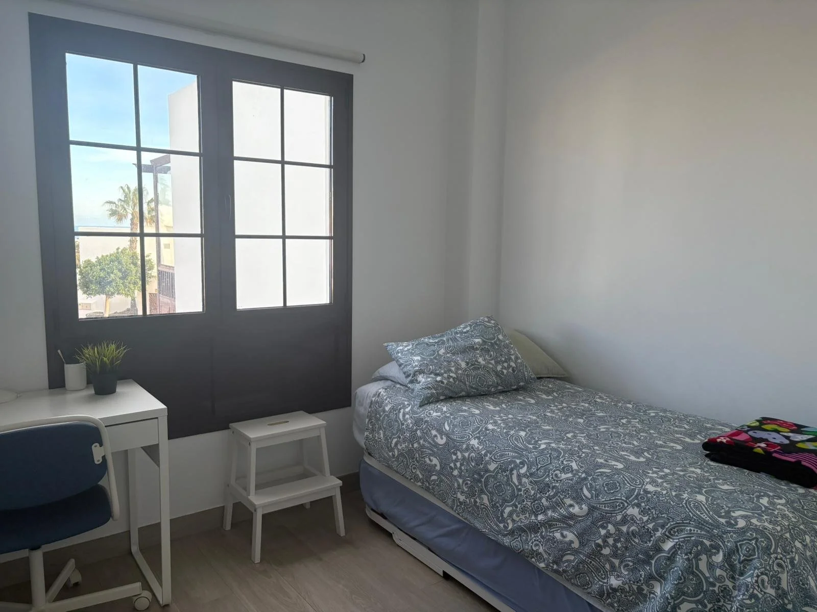 Minimalist bedroom with a single bed, patterned bedspread, desk, chair, and window.