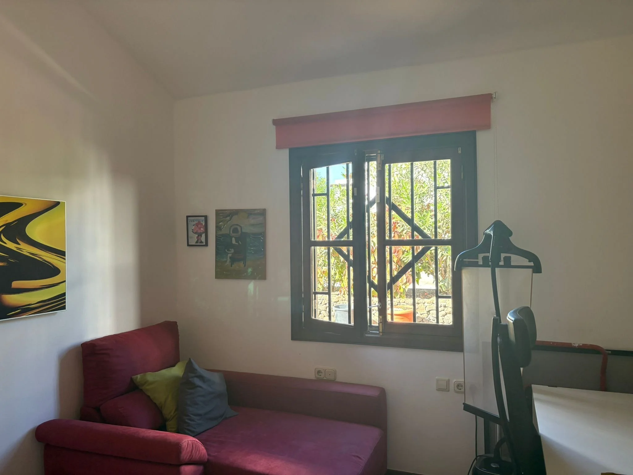 A cozy room with a purple sofa and decorative pillows, artwork on the walls, a wooden-framed window with a view outside, and a clothes steamer beside a desk.