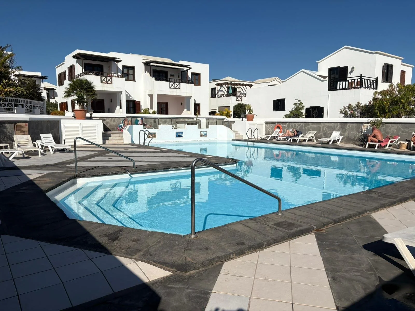 Swimming pool with lounge chairs, white buildings, and blue sky in a resort setting.