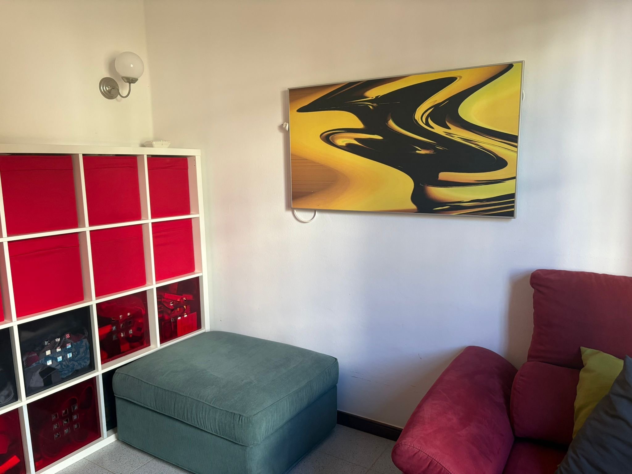 Room with abstract art, red sofa, green ottoman, storage unit with red bins, and wall lamp.