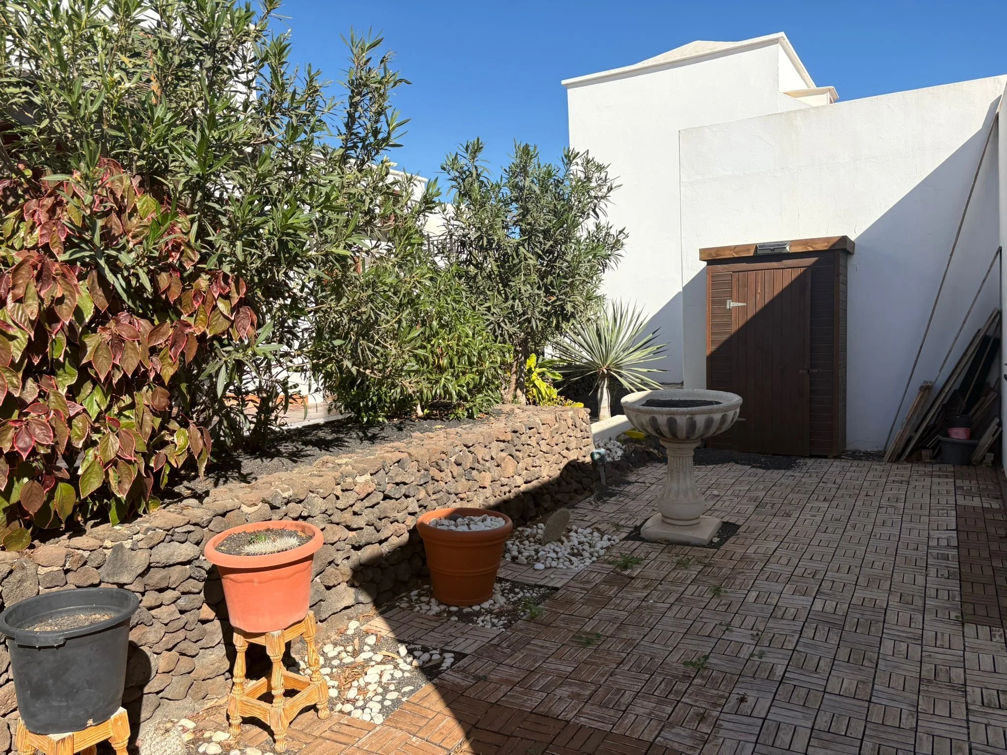 Outdoor patio area with stone wall, potted plants, pedestal birdbath, and small wooden shed.