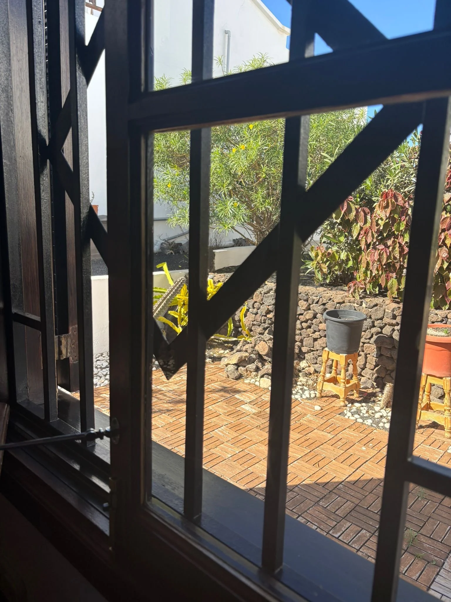 view through window with iron bars overlooking a garden with brick patio, potted plants, and a stone wall