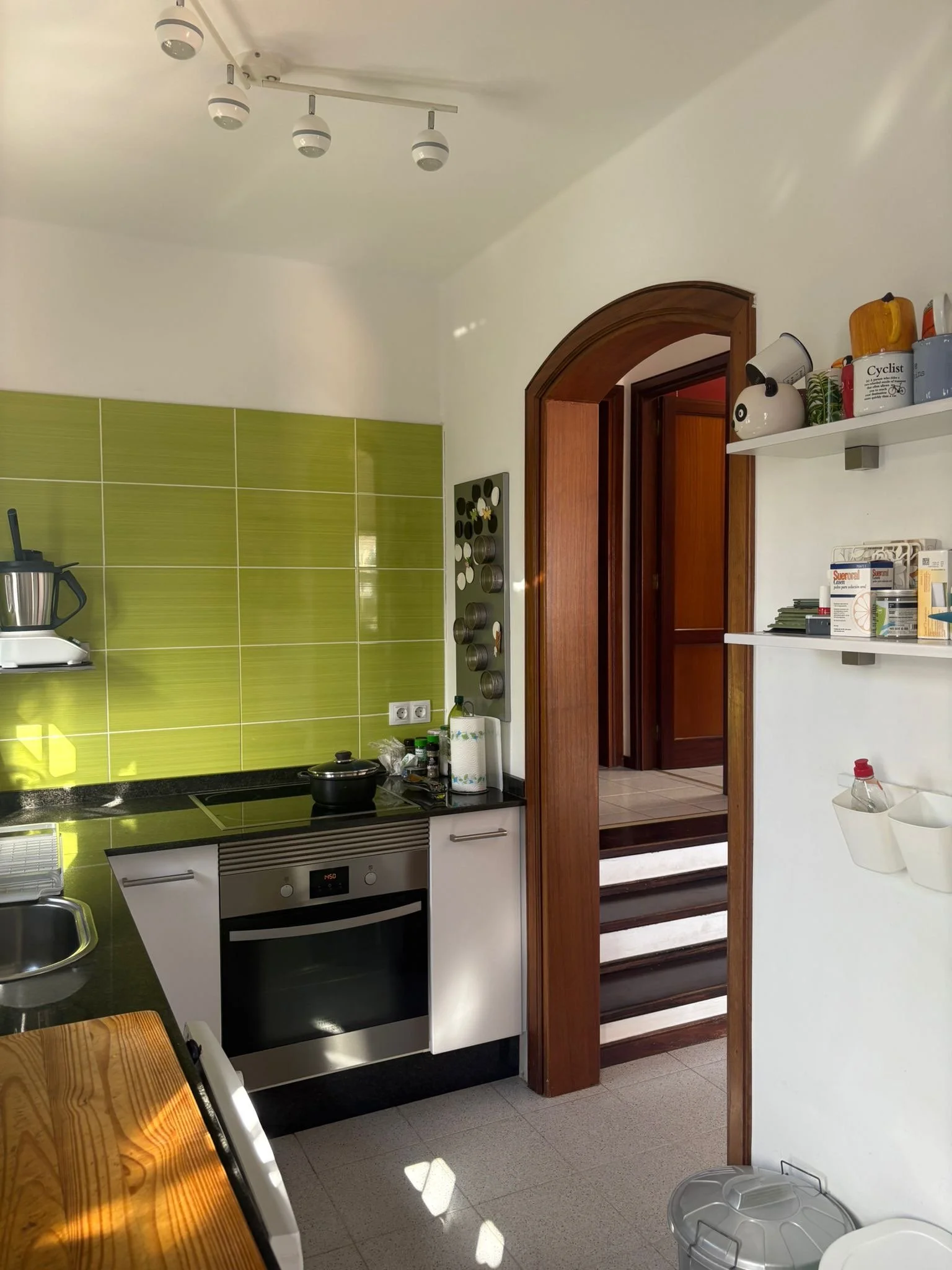Modern kitchen with green tile backsplash, electric stove, oven, and stainless steel sink. Wooden dining table in foreground, wall shelves with kitchen items, and a doorway leading to another room.