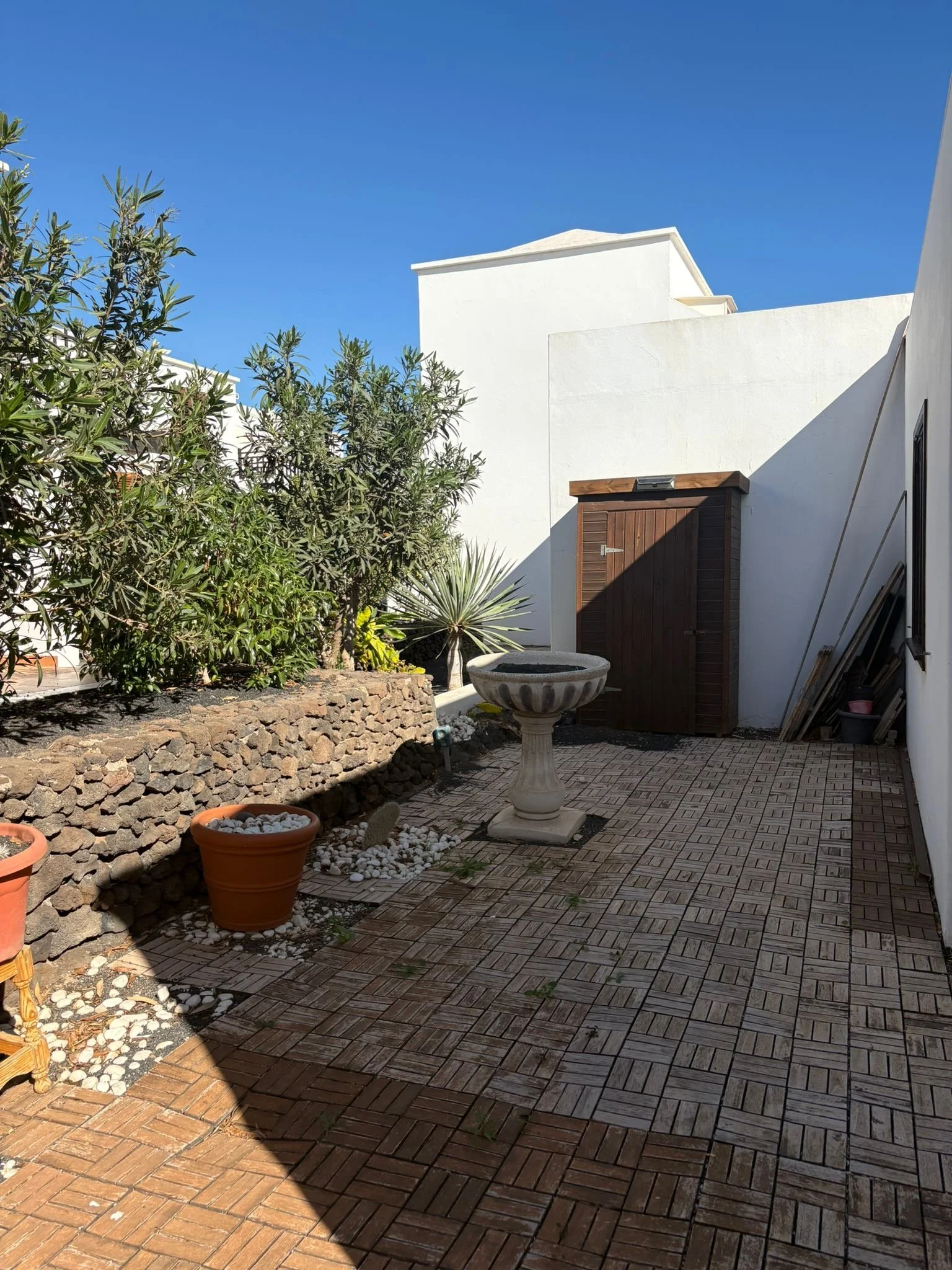 Outdoor patio with stone tiles, plant pots, a bird bath, and greenery against a white building wall.