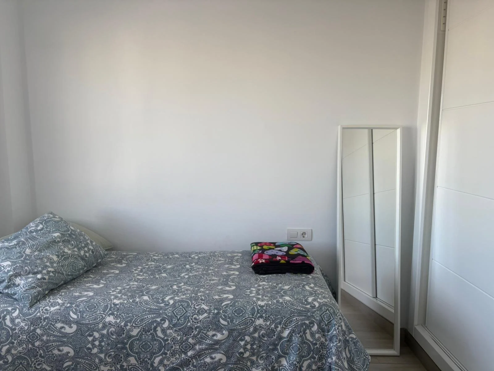 Minimalist bedroom with a single bed featuring a blue patterned duvet and pillow, folded colorful blanket, and a full-length mirror propped against the wall.