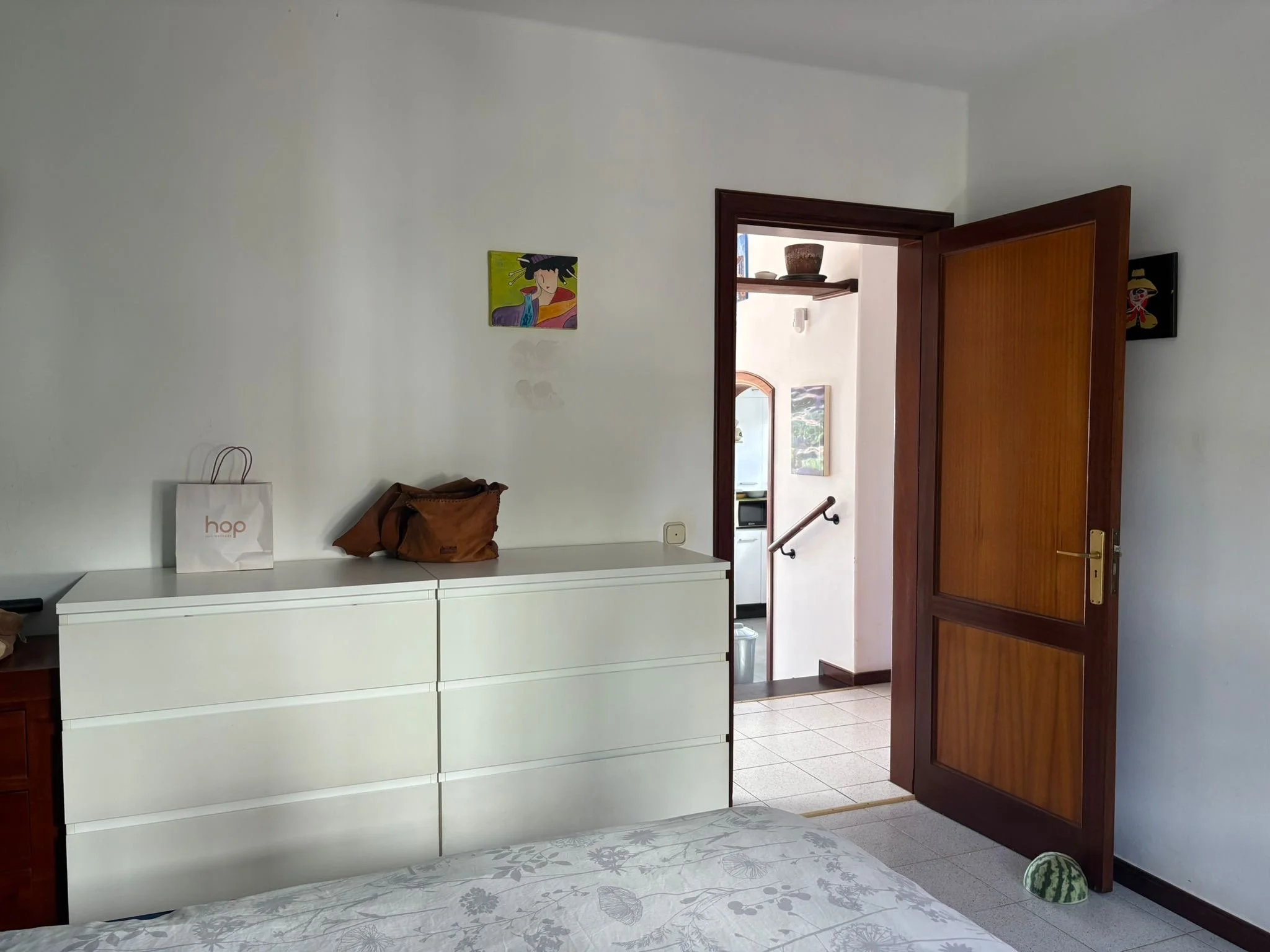 Interior of a bedroom with a white dresser, a bag with "hop" logo, brown bag, open wooden door leading to a hallway, wall art featuring colorful portraits, and a bed with patterned bedding.