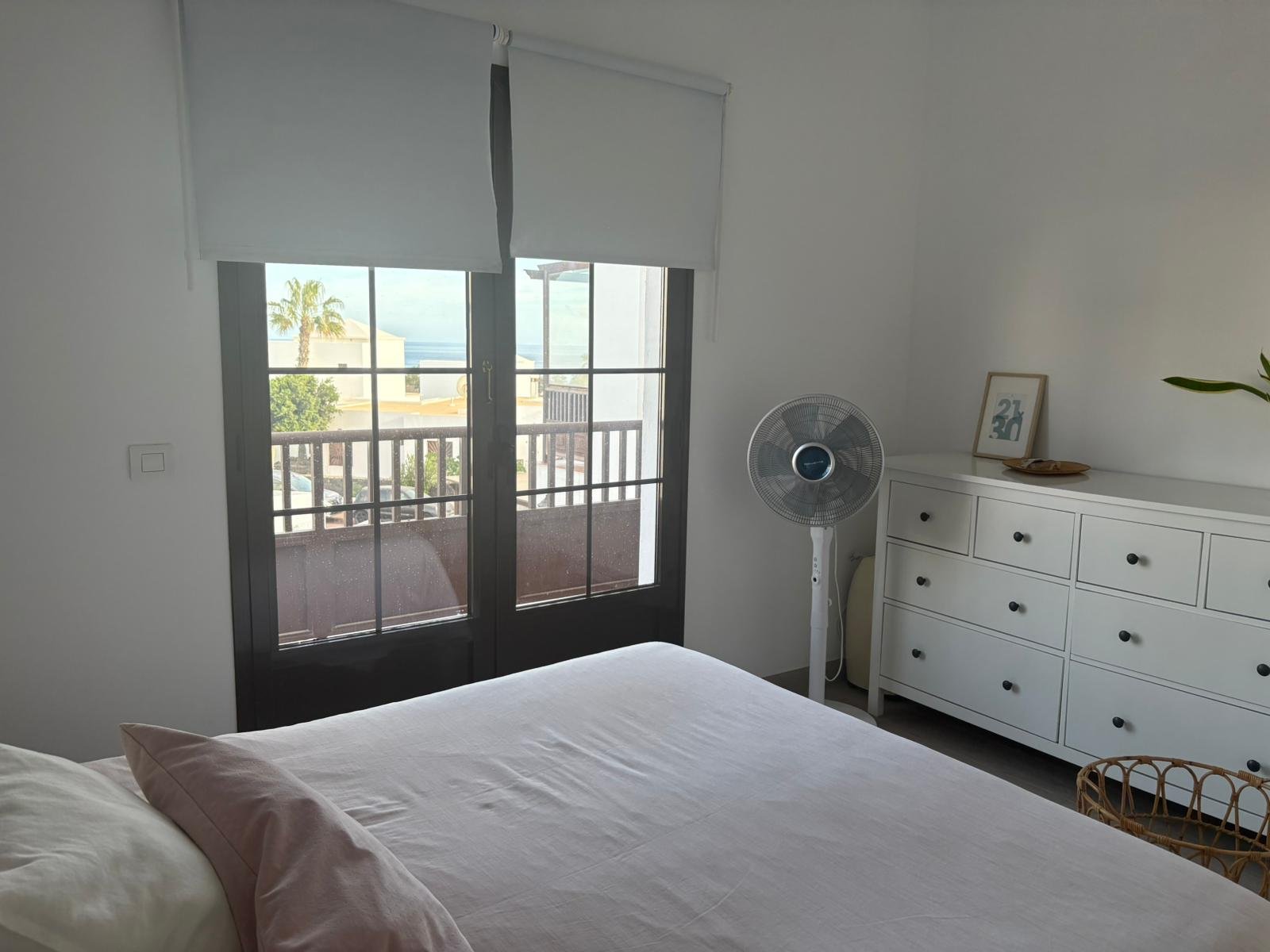 Bedroom with a bed, white dresser, standing fan, and a view through a glass door leading to a balcony with a palm tree and ocean in the background.